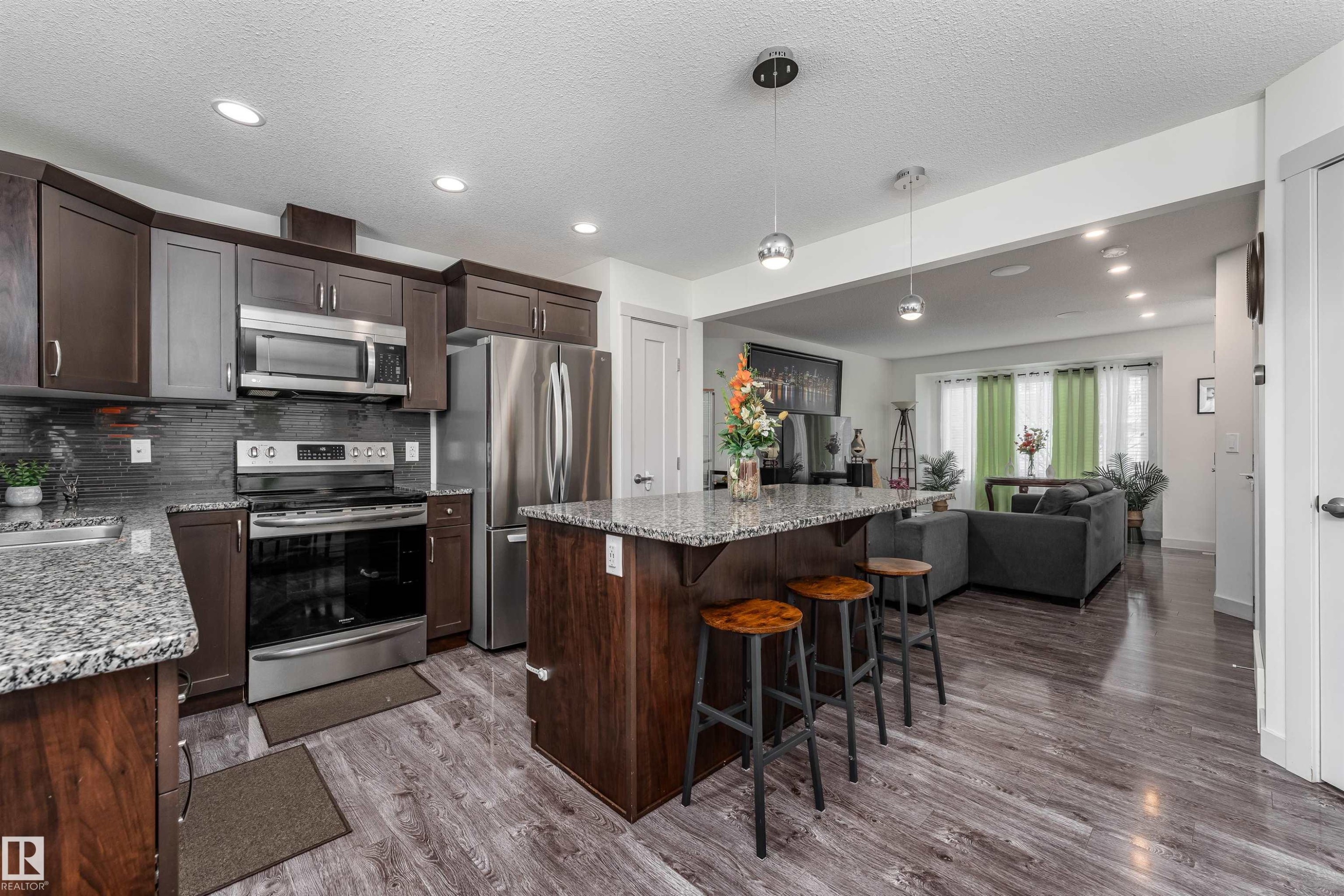 Kitchen featuring wood-finish flooring, stainless steel appliances, dark wood cabinetry, a tiled backsplash, and an island with a stone countertop and pendant lighting - 15 5203 149 Avenue, Edmonton, AB - Indoor Photo Showing Kitchen With Upgraded Kitchen