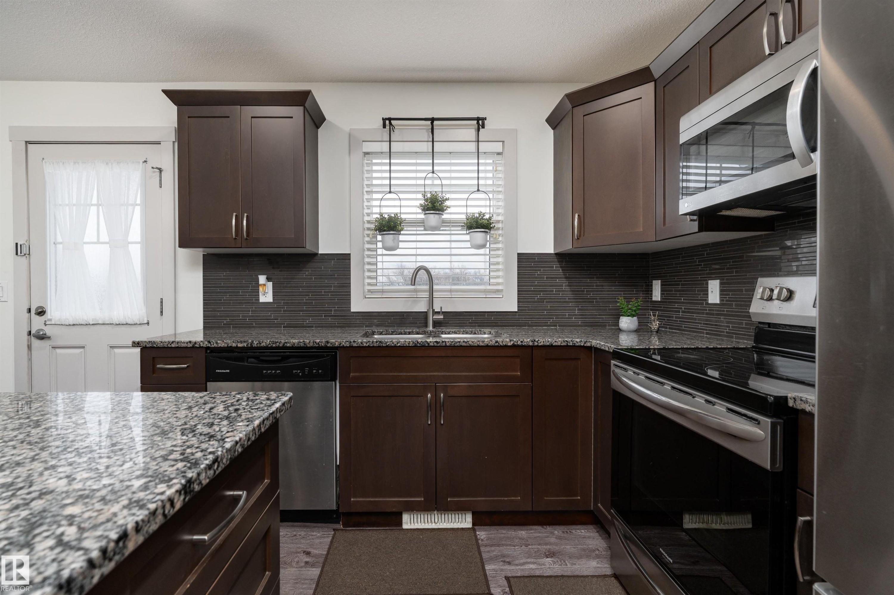 Kitchen featuring dark wood cabinetry, granite countertops, a dark tile backsplash, stainless steel appliances, and wood-finish flooring - 15 5203 149 Avenue, Edmonton, AB - Indoor Photo Showing Kitchen With Upgraded Kitchen