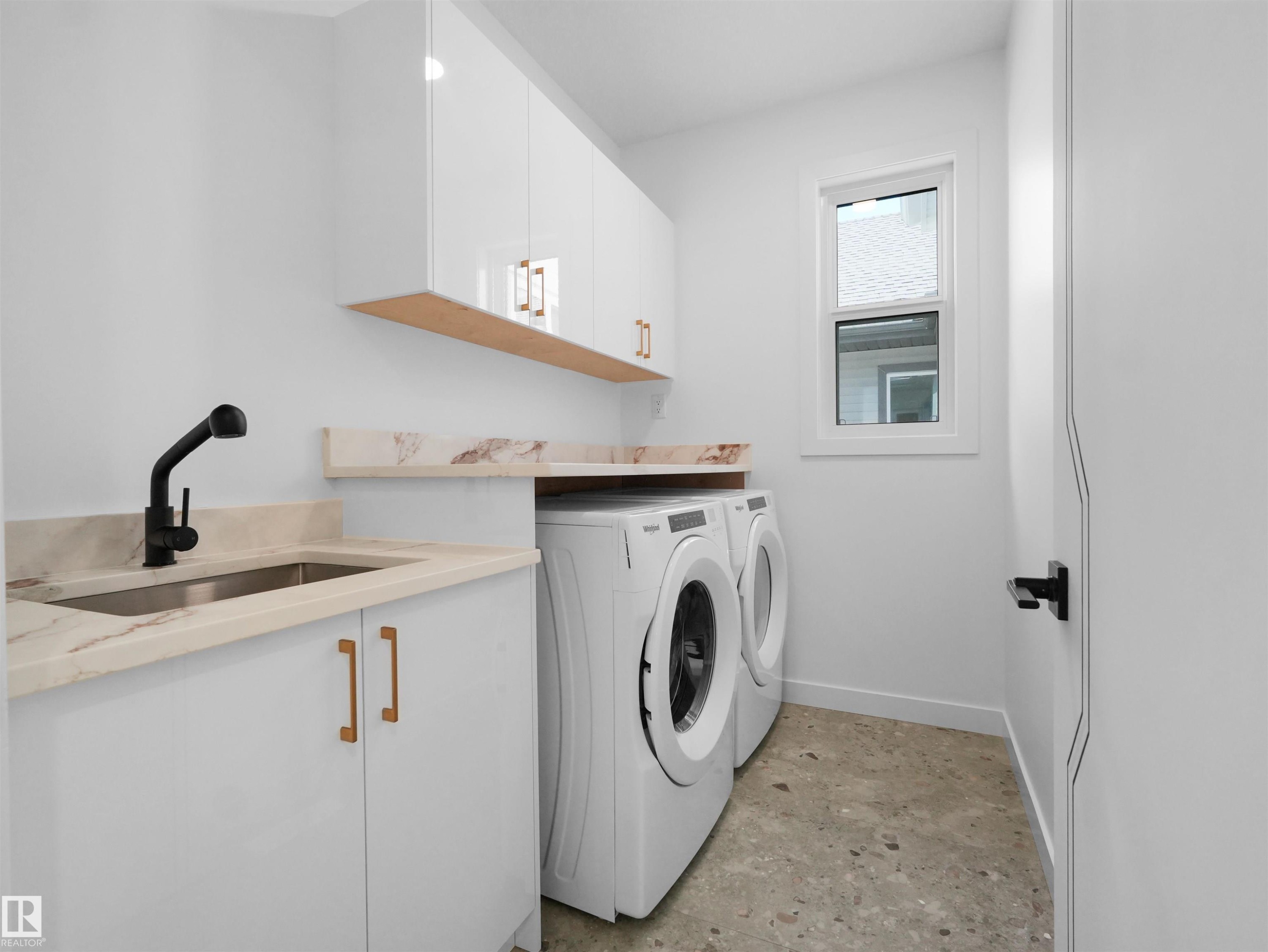 Dedicated laundry area featuring a stainless steel sink with matte black faucet, light stone-finish countertops, white cabinetry with gold-tone hardware, and a window - 2070 Collip Crescent Sw, Edmonton, AB - Indoor Photo Showing Laundry Room