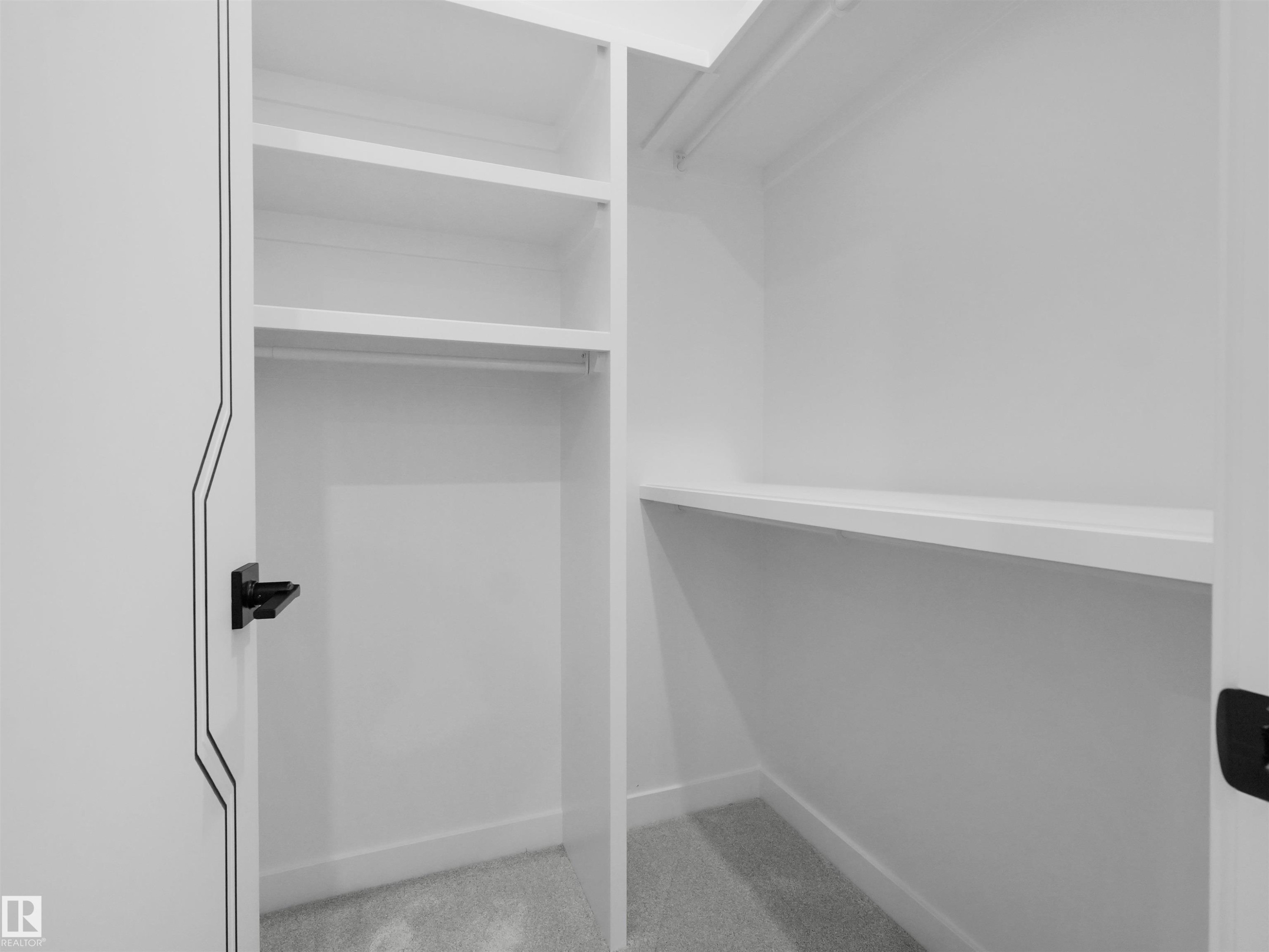 Custom closet system with multiple white shelves and hanging rods, plush gray carpeting, white baseboards, and a modern black door handle - 2070 Collip Crescent Sw, Edmonton, AB - Indoor With Storage