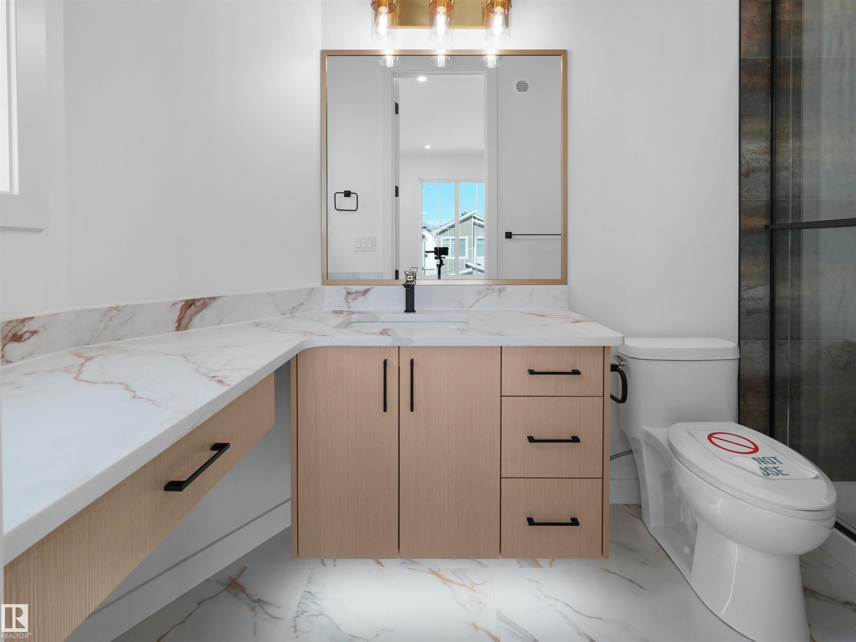 Bathroom featuring an expansive white and reddish-brown veined countertop, a light wood-finish vanity with ample storage, a brass-toned light fixture, a mirrored medicine cabinet, and white flooring with similar veining - 2070 Collip Crescent Sw, Edmonton, AB - Indoor Photo Showing Bathroom