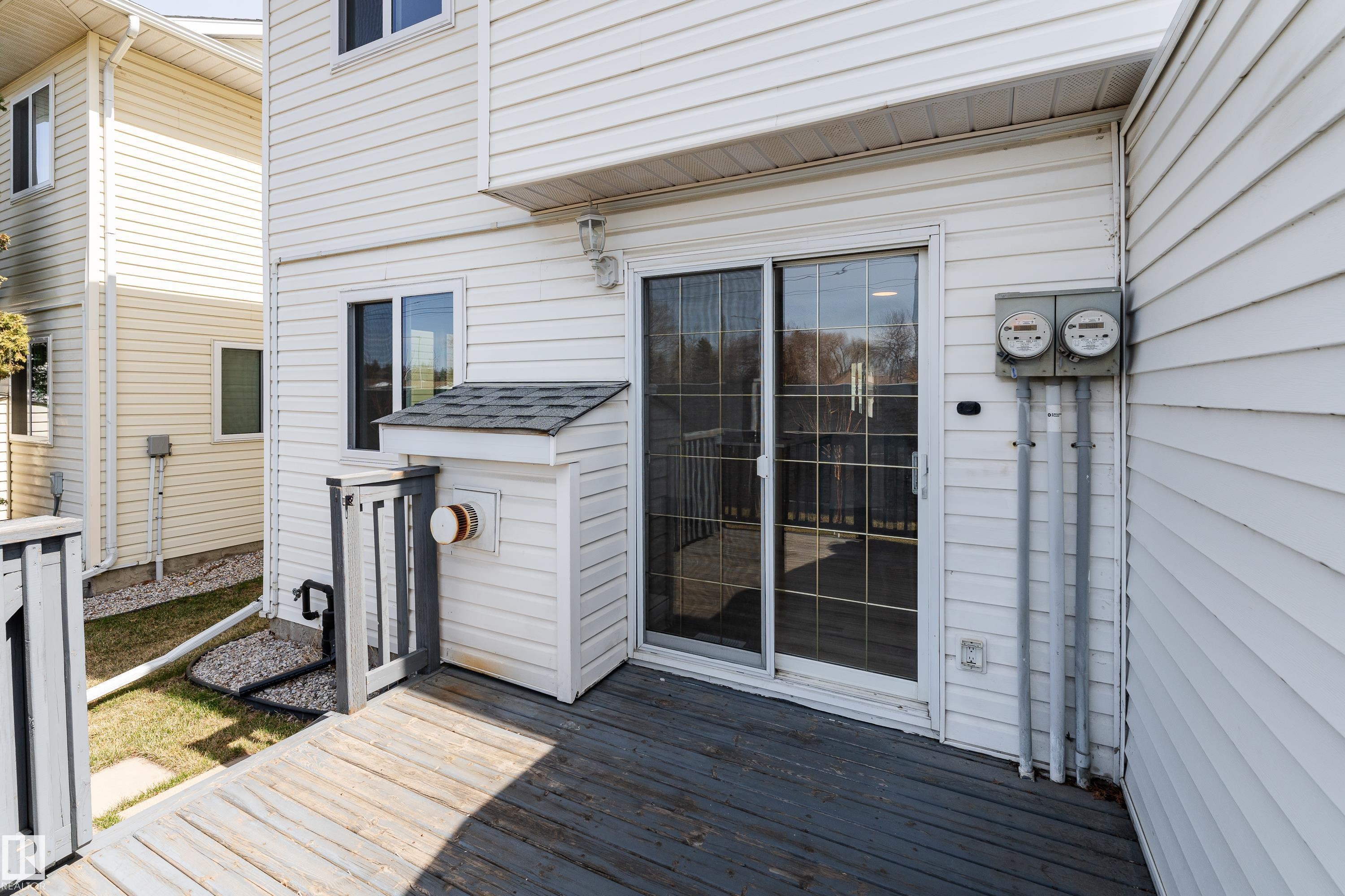 Private deck area featuring white vinyl siding, a sliding glass door, a window with dark trim, and a white utility enclosure with a shingled roof - #63 1033 Youville Dr W Nw, Edmonton, AB - Outdoor With Deck Patio Veranda With Exterior