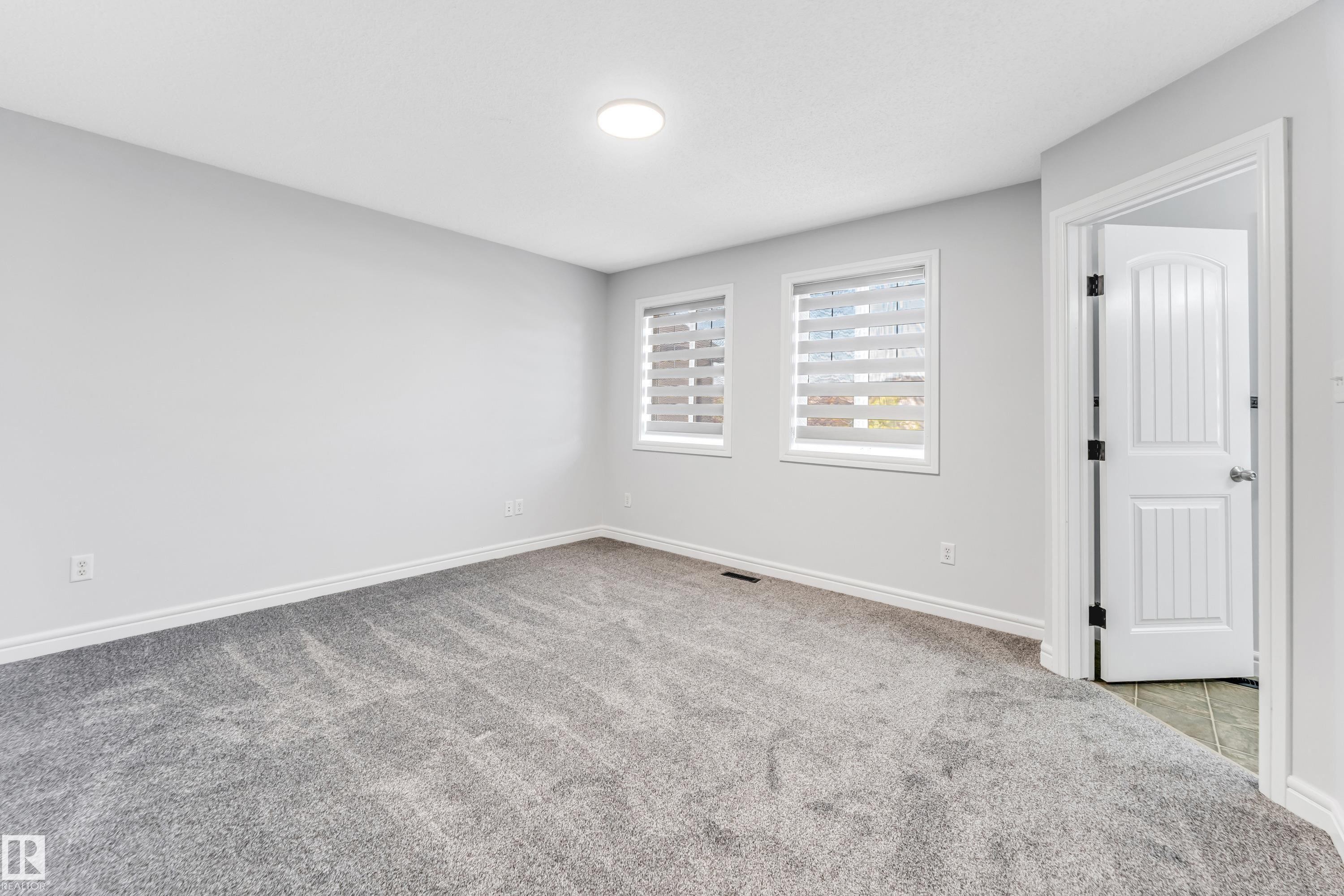 Light gray carpeted room with light gray walls, two windows featuring horizontal blinds, and a white six-panel interior door - 1921 119A Street, Edmonton, AB