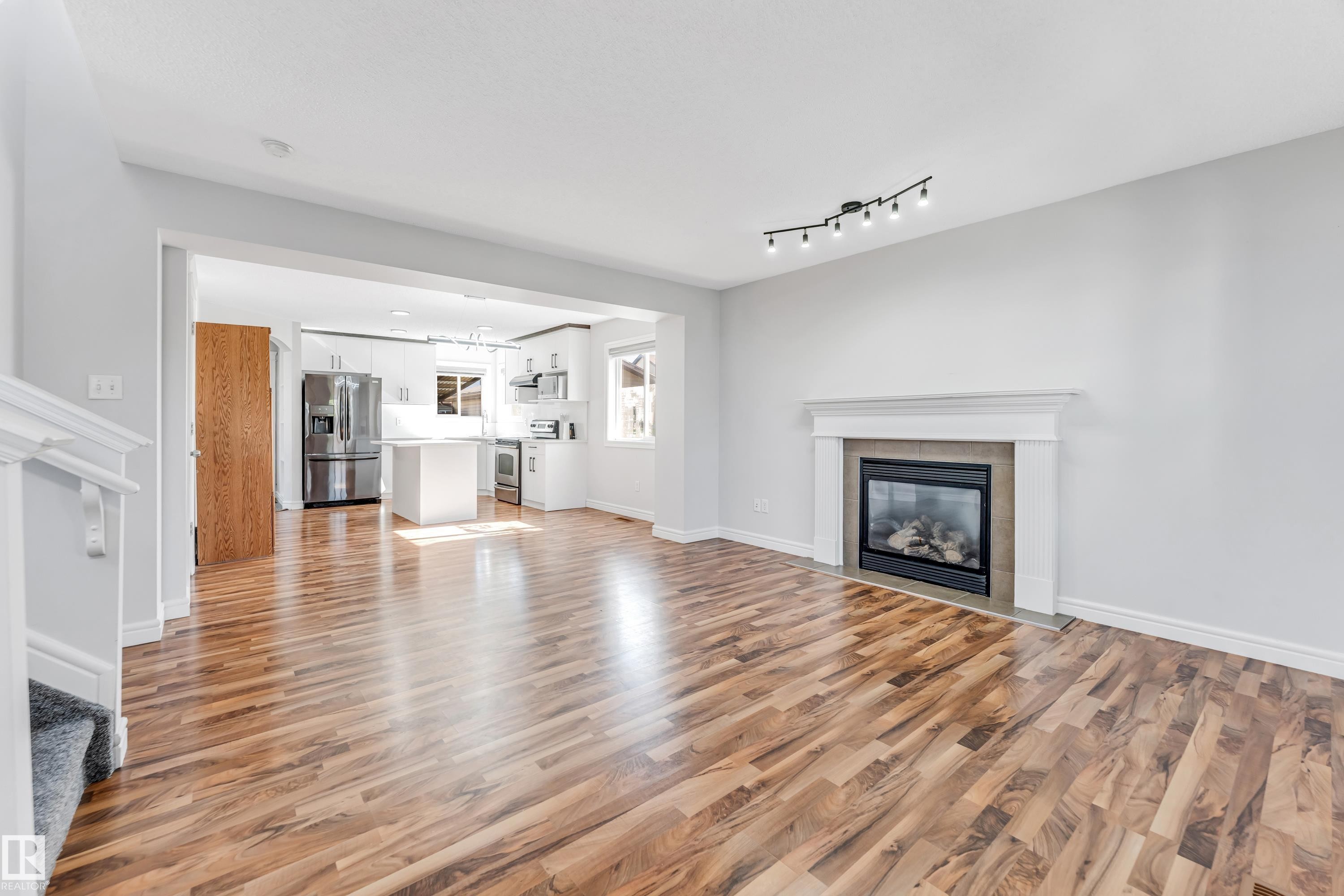 Open-concept living area featuring wood-finish flooring, a contemporary fireplace with a white mantel, and track lighting - 1921 119A Street, Edmonton, AB