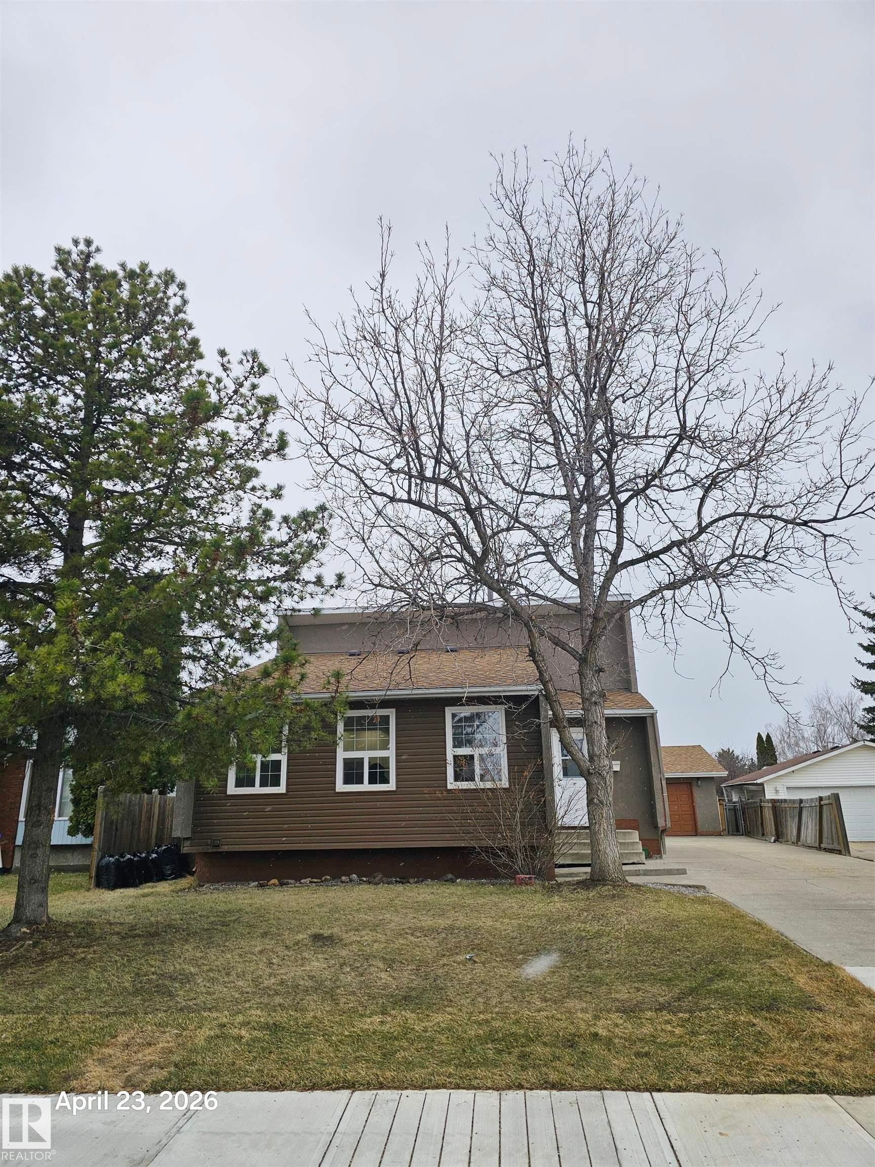 Brown siding exterior with white window frames, a gray shingle roof, mature front yard trees, and a concrete driveway - 20 Hunt Road, Edmonton, AB - Outdoor
