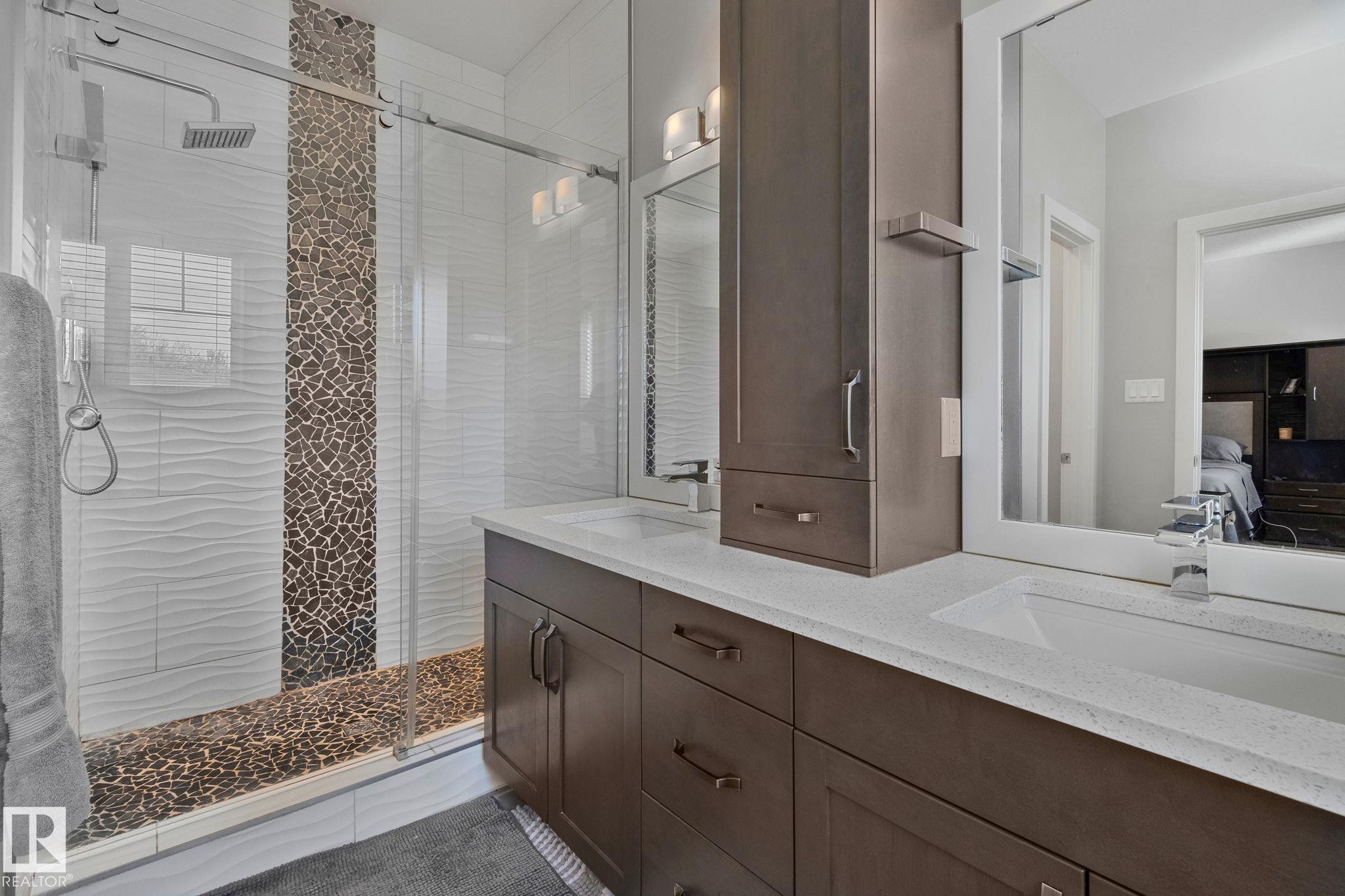 Contemporary bathroom featuring a glass-enclosed shower with pebble-style flooring and textured white wall tile, dual vanity with integrated sinks and white countertops, dark wood-finish cabinetry, and a tall built-in storage cabinet - 67 Enchanted Way Nw, St. Albert, AB - Indoor Photo Showing Bathroom