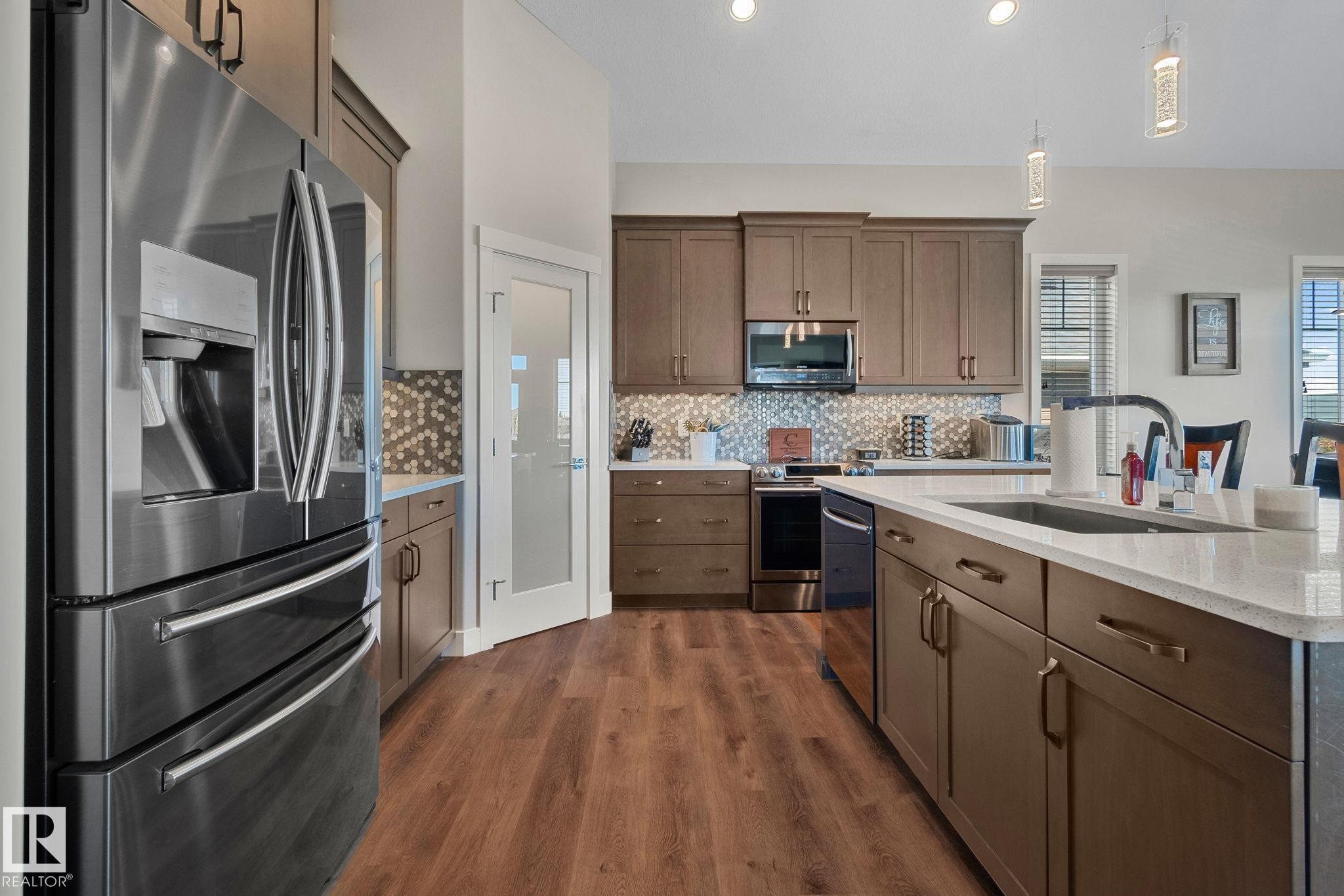 Contemporary kitchen featuring wood-finish flooring, stainless steel appliances, a large island with an undermount sink, mosaic tile backsplash, and recessed lighting - 67 Enchanted Way Nw, St. Albert, AB - Indoor Photo Showing Kitchen With Upgraded Kitchen