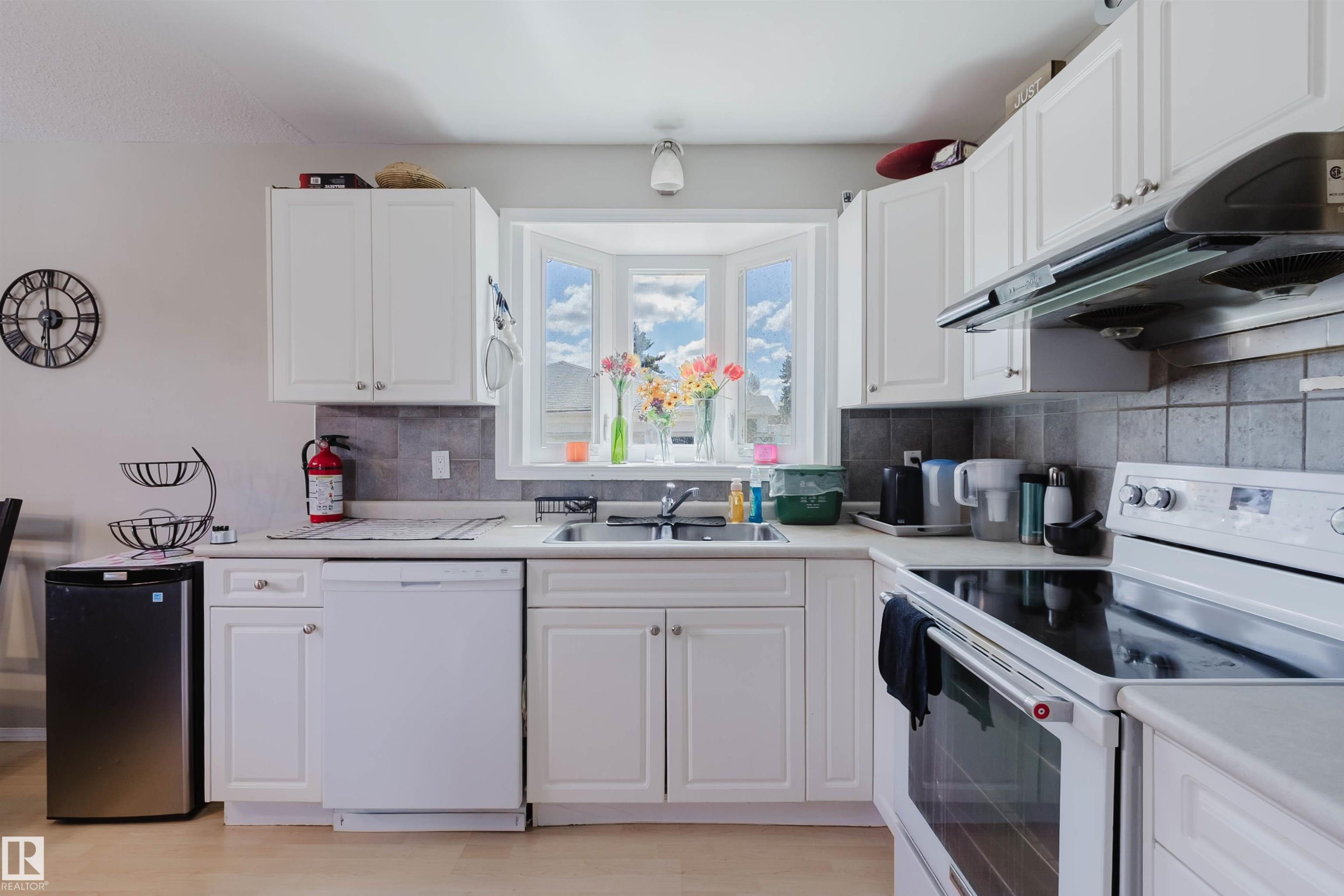 Kitchen featuring white cabinetry, a bay window, stainless steel sink, tile backsplash, and wood-finish flooring - 10519 36A Avenue, Edmonton, AB - Indoor Photo Showing Kitchen With Double Sink