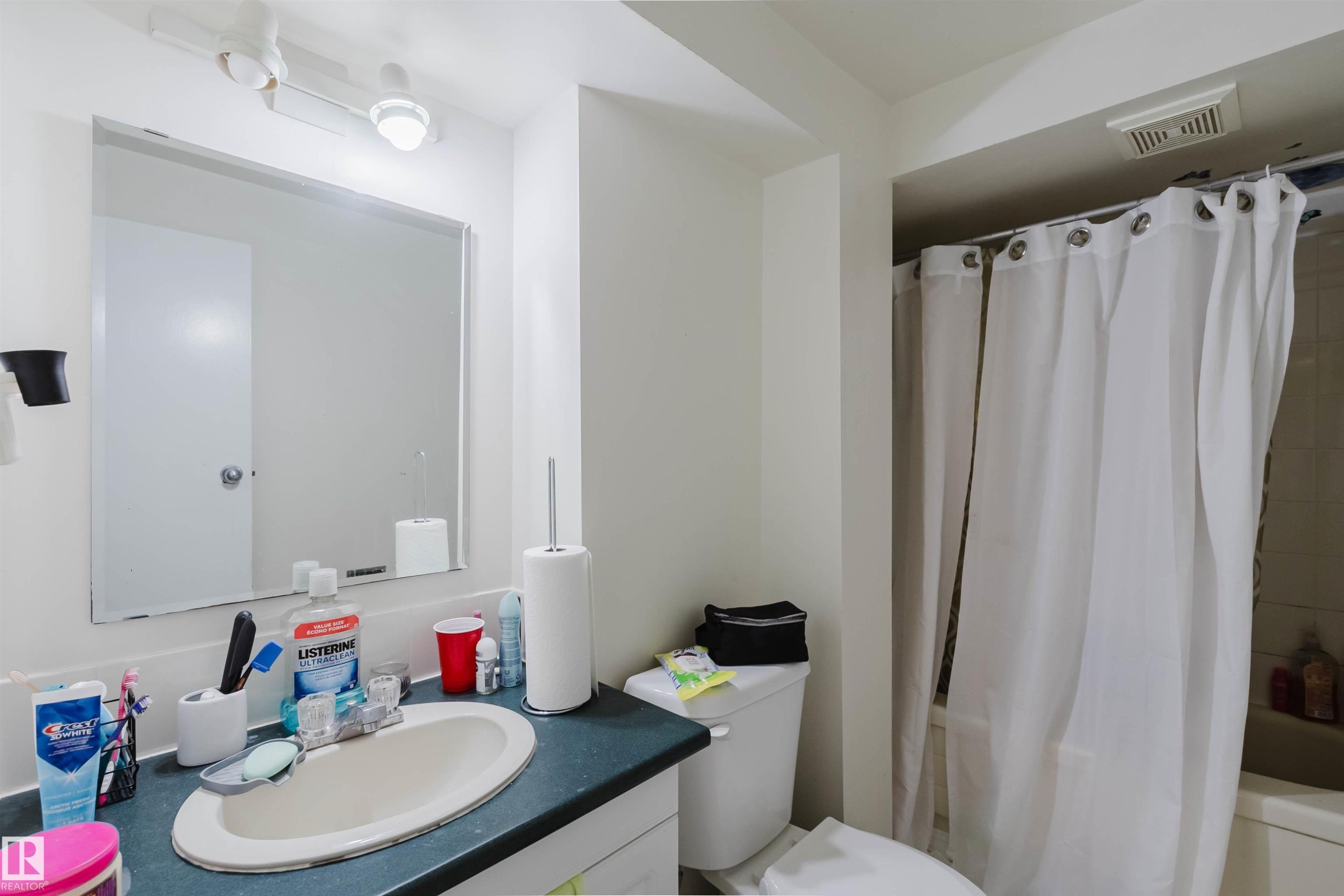 Bathroom featuring a track light fixture, a wall-mounted mirror, a vanity with an integrated sink and dark countertop, a toilet, and a shower-over-tub combination with a shower curtain rod - 10519 36A Avenue, Edmonton, AB - Indoor Photo Showing Bathroom