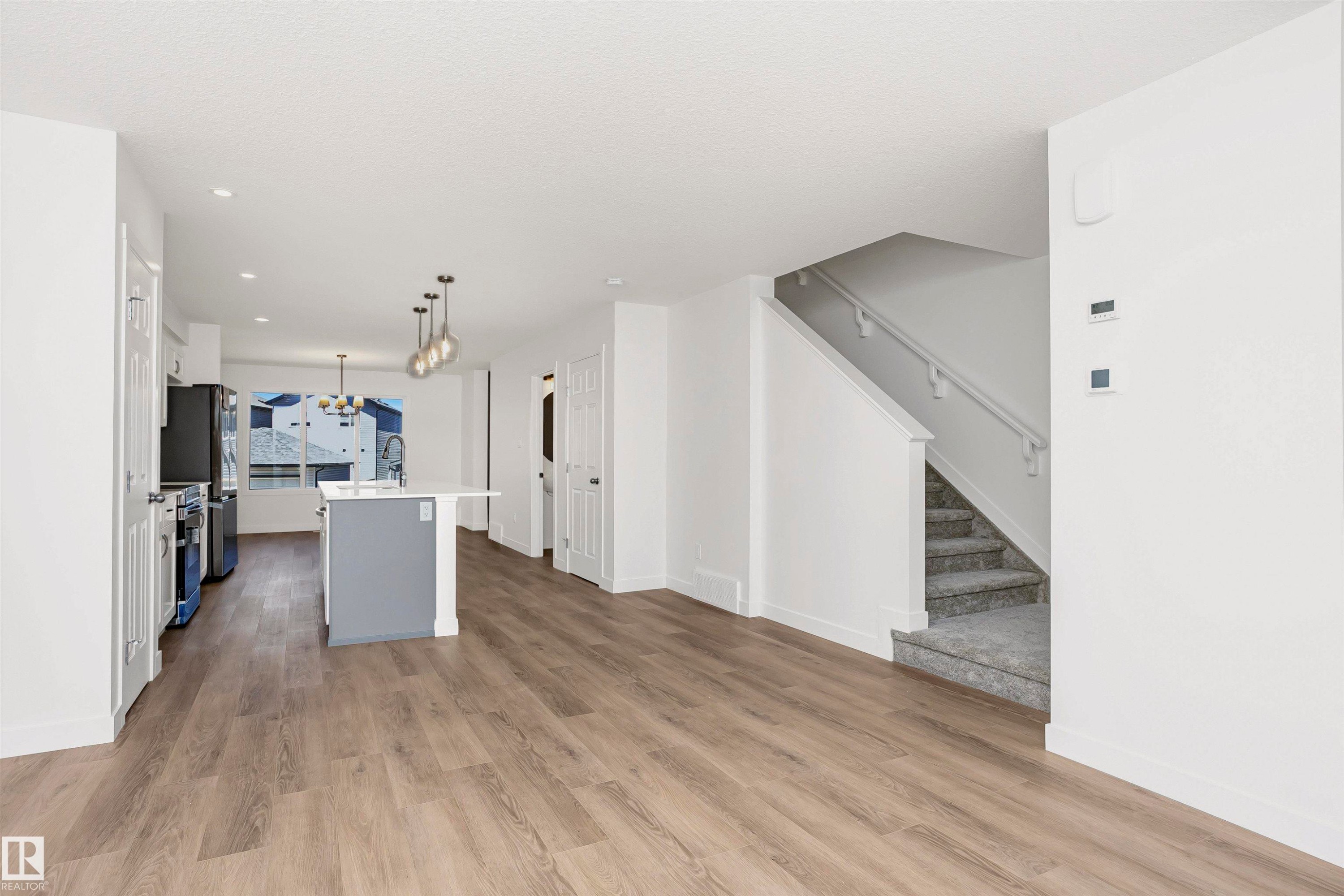 Spacious open-concept living area featuring wood-finish flooring, a kitchen island with seating, pendant lighting, and a staircase with carpeting and a handrail - 9552 Carson Bend Bend, Edmonton, AB - Indoor