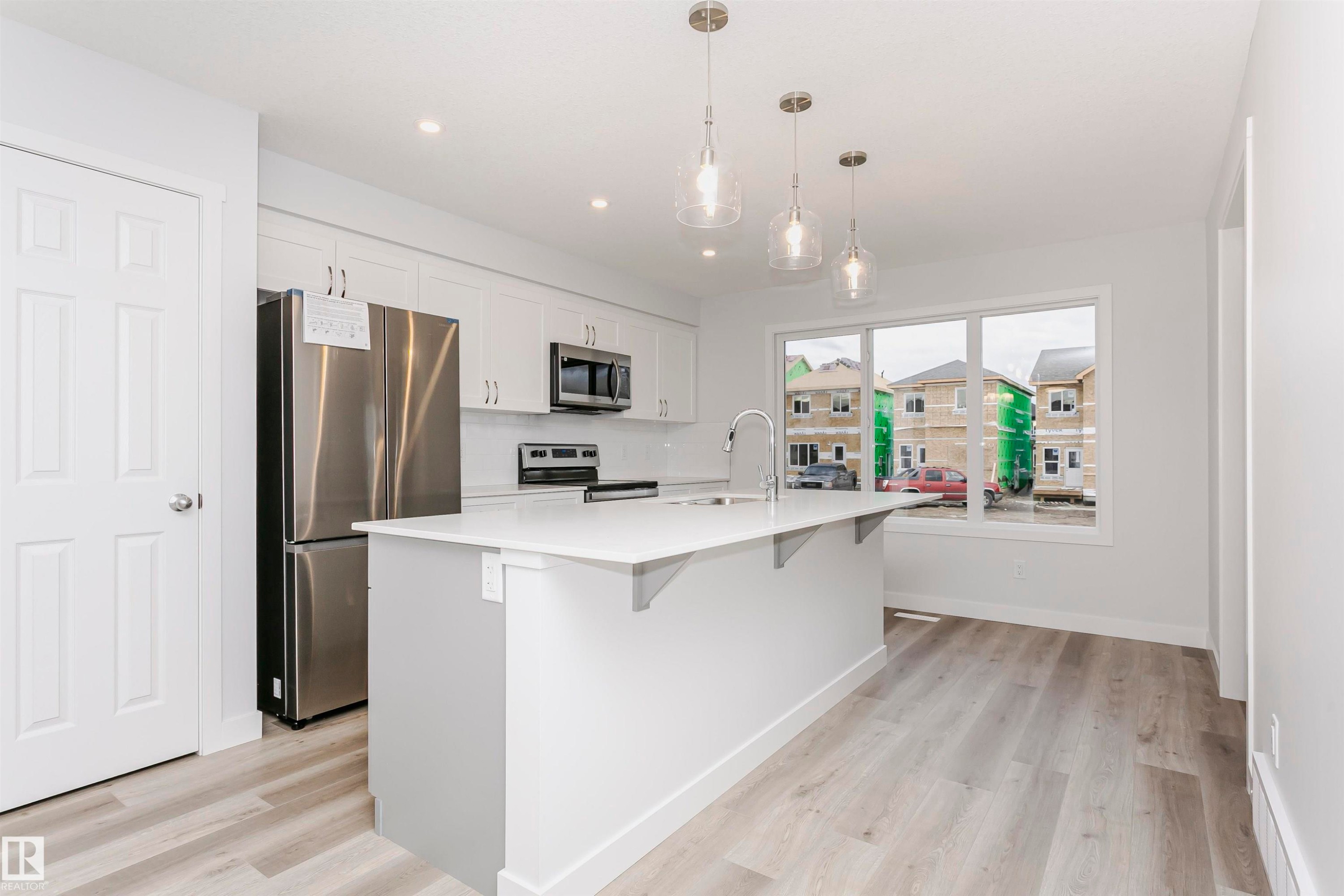 Modern kitchen featuring a central island with a sink, white cabinetry, stainless steel appliances, pendant lighting, and wood-finish flooring - 9564 Carson Bend Bend, Edmonton, AB - Indoor Photo Showing Kitchen With Upgraded Kitchen