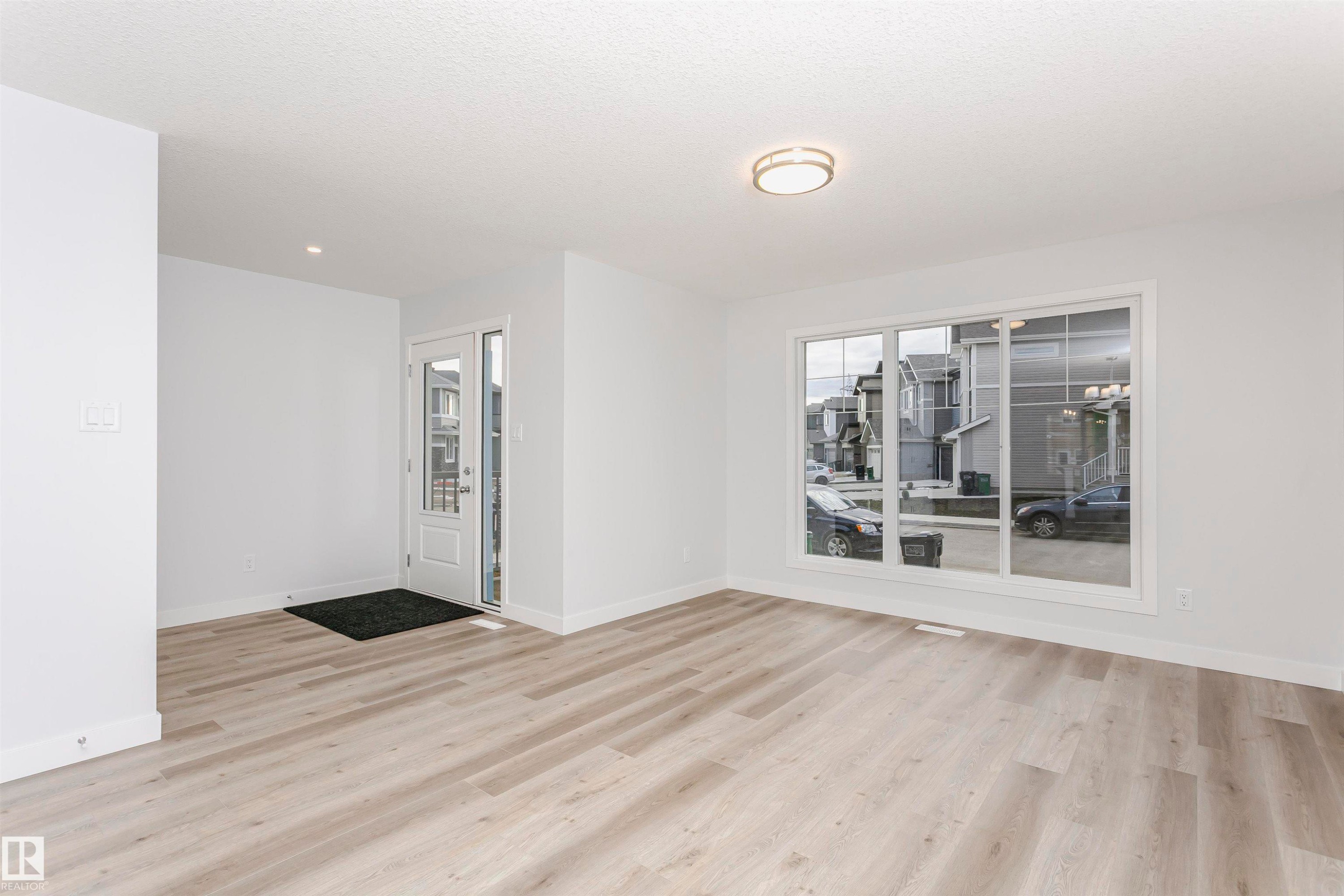 Bright interior space featuring a light wood-finish flooring, white walls, and a large front-facing window - 9564 Carson Bend Bend, Edmonton, AB - Indoor
