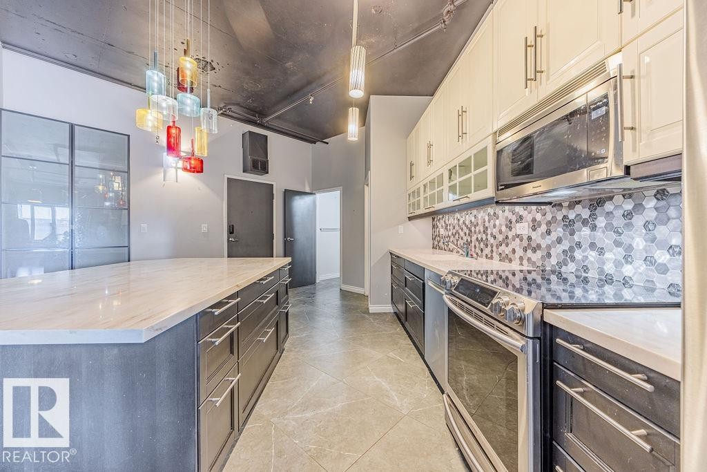 Kitchen featuring an expansive island with a light-toned countertop, stainless steel appliances, two-tone cabinetry, a hexagonal tile backsplash, and a multi-colored pendant light fixture - 303 10179 105 Street, Edmonton, AB