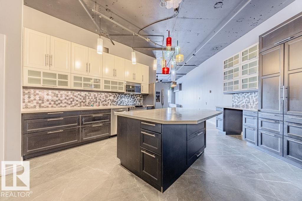 Kitchen featuring a central island with a light-toned countertop, dark wood-finish cabinetry, a hexagonal tile backsplash, and a contemporary multi-colored pendant light fixture - 303 10179 105 Street, Edmonton, AB