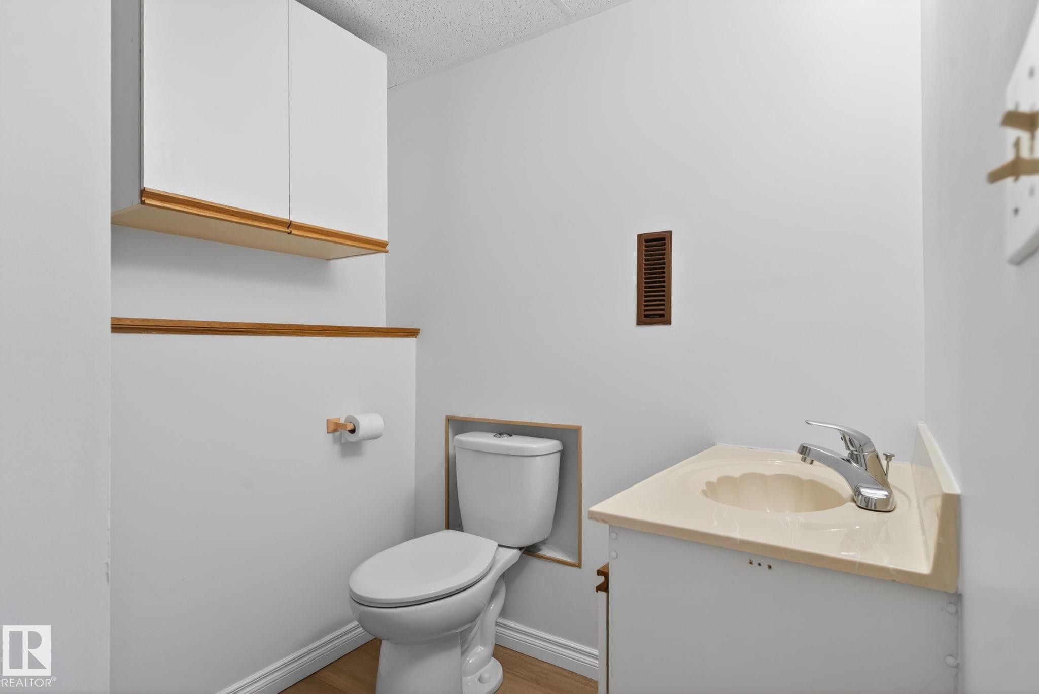 Bathroom featuring a white toilet, wood-finish flooring, a white vanity with an integrated sink, chrome faucet, and overhead white cabinetry with wood trim - 8403 190 Street, Edmonton, AB - Indoor Photo Showing Bathroom