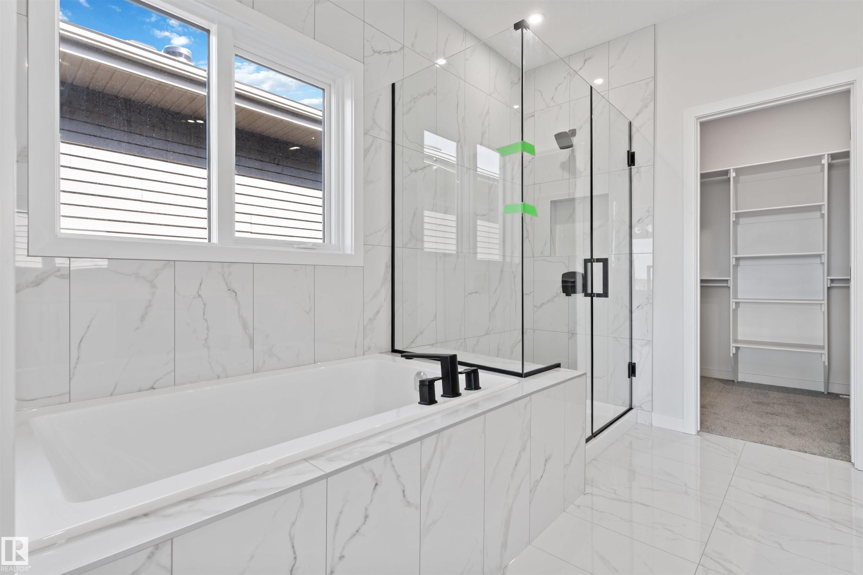 Full bathroom featuring a rectangular soaking tub, glass-enclosed shower with black fixtures, white marble-look tile flooring, and a walk-in closet with built-in shelving - 1849 Crossbill Loop Nw, Edmonton, AB - Indoor