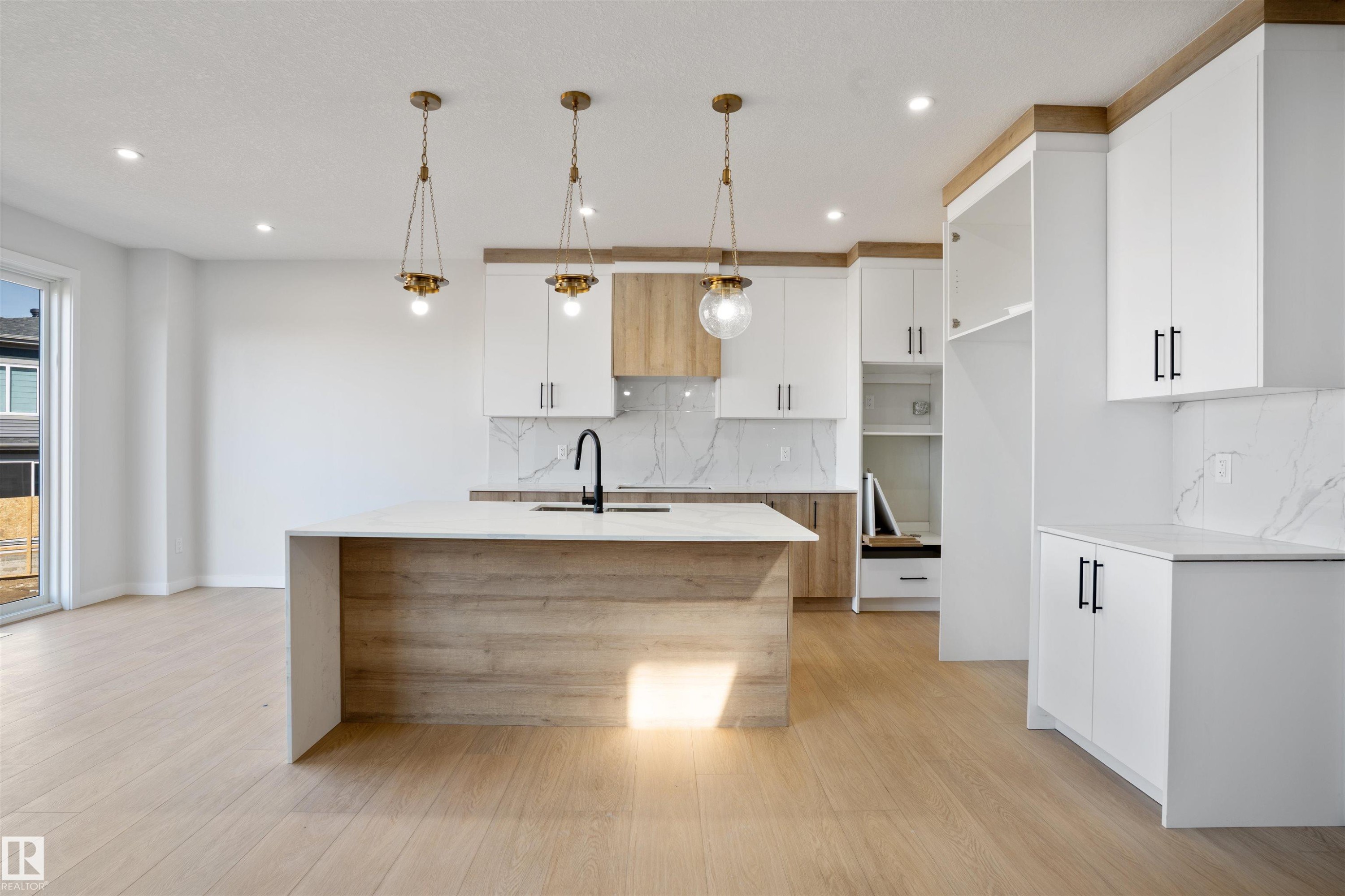 Modern kitchen featuring a large central island with a waterfall countertop, light wood-finish cabinetry, and white overhead cabinets with matte black hardware - 1849 Crossbill Loop Nw, Edmonton, AB - Indoor Photo Showing Kitchen