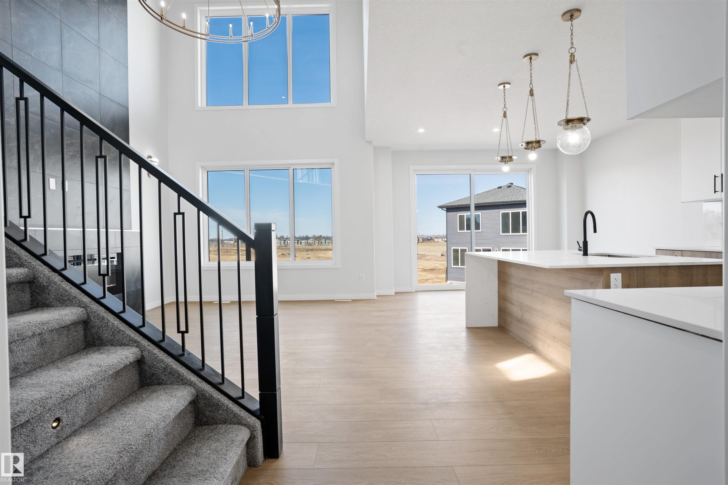 Two-story living space featuring an open-concept design with light wood-finish flooring, a contemporary staircase with carpeted treads and black metal railings, and a kitchen island with a white countertop and wood-finish paneling - 1849 Crossbill Loop Nw, Edmonton, AB - Indoor