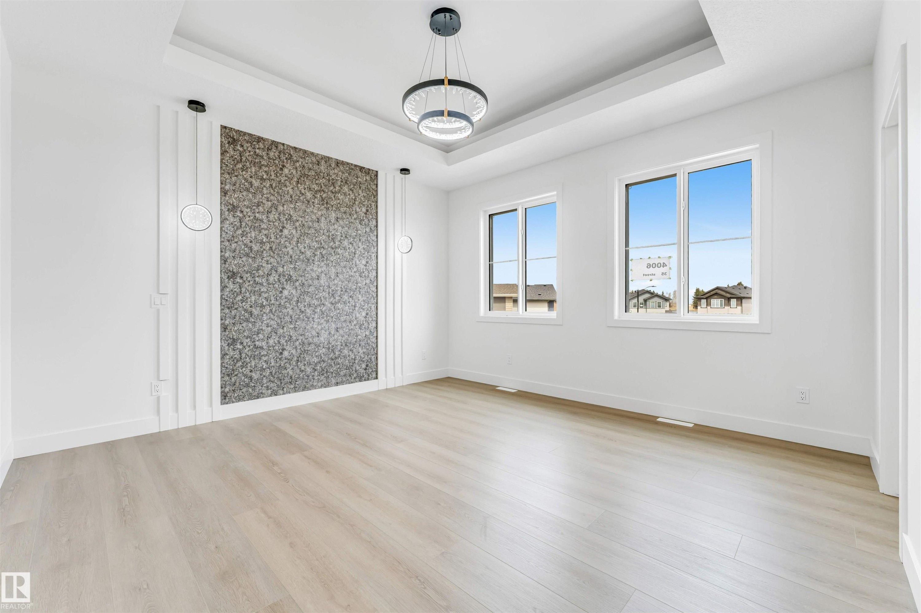 Spacious room featuring a textured accent wall with decorative paneling and integrated circular sconces, wood-finish flooring, a tray ceiling with a contemporary light fixture, and two large windows providing natural light - 4006 35 St, Beaumont, AB - Indoor Photo Showing Other Room