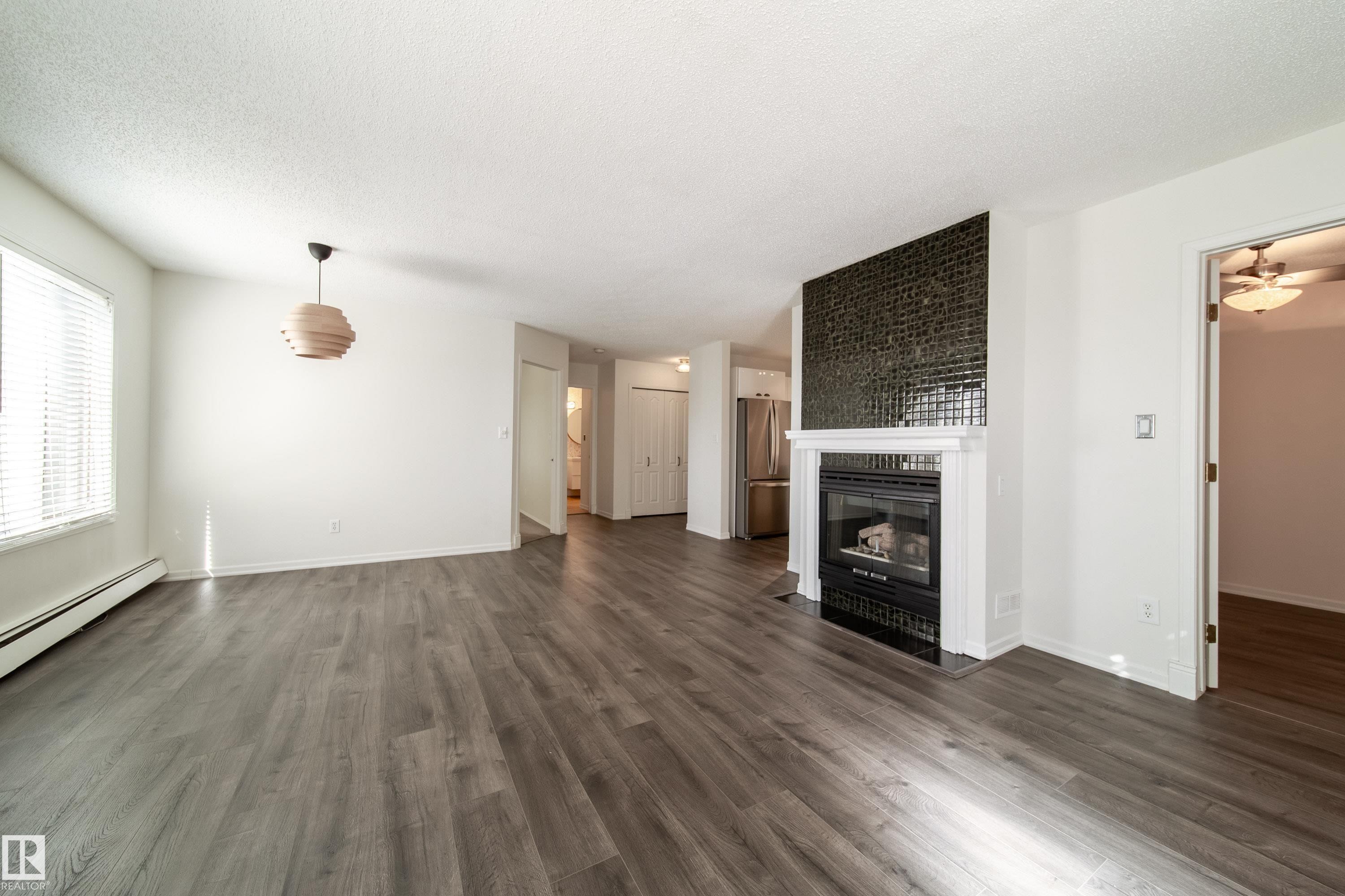Spacious living area featuring wood-finish flooring, a contemporary fireplace with a decorative tile surround and white mantel, and a large window with blinds - 302 9138 83 Avenue, Edmonton, AB - Indoor Photo Showing Living Room With Fireplace