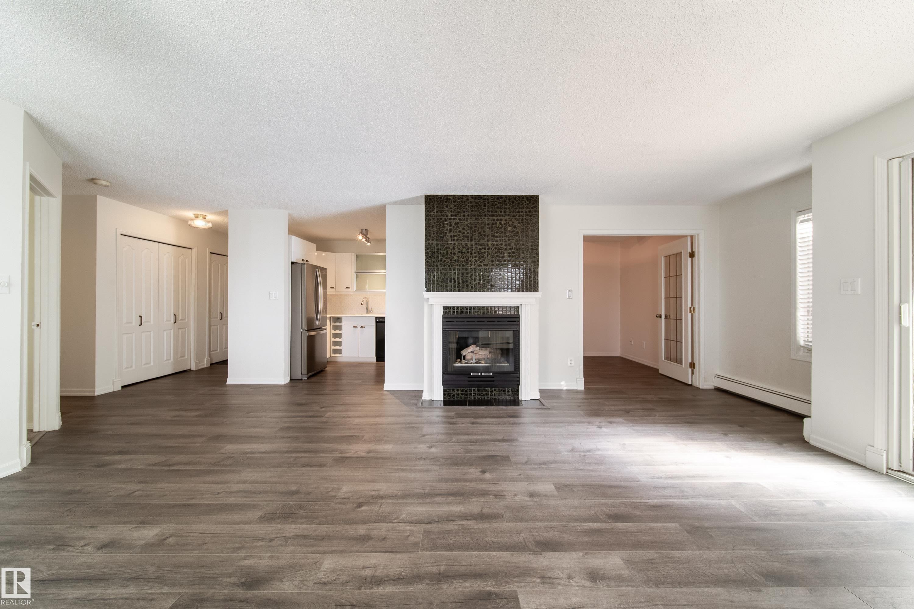 Spacious living area featuring wood-finish flooring, a fireplace with a dark mosaic tile surround and white mantel, and ample natural light from the sliding glass door - 302 9138 83 Avenue, Edmonton, AB - Indoor Photo Showing Living Room With Fireplace