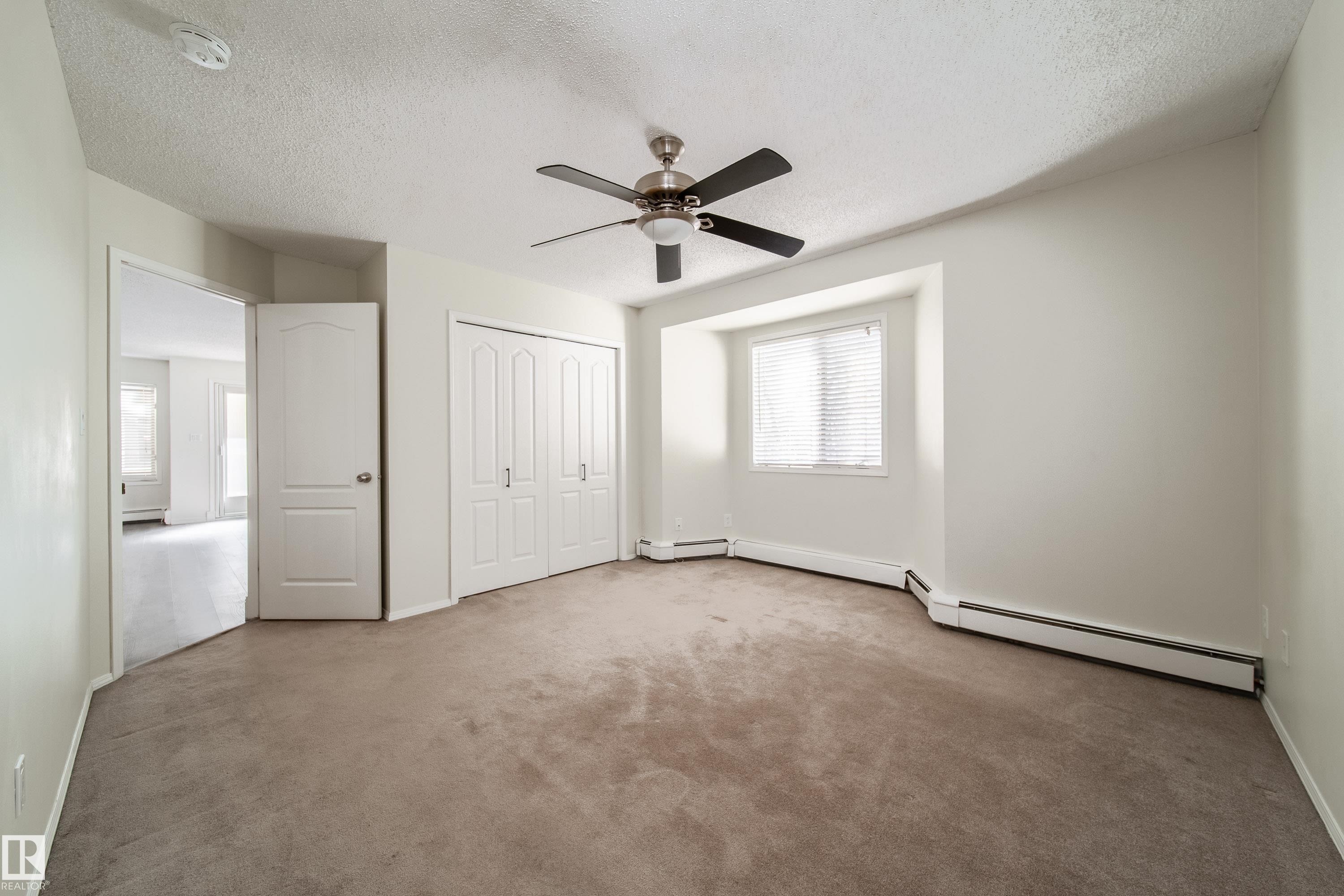 Carpeted room featuring a ceiling fan with light fixture, window with horizontal blinds, bi-fold closet doors, and baseboard heating - 302 9138 83 Avenue, Edmonton, AB - Indoor Photo Showing Other Room
