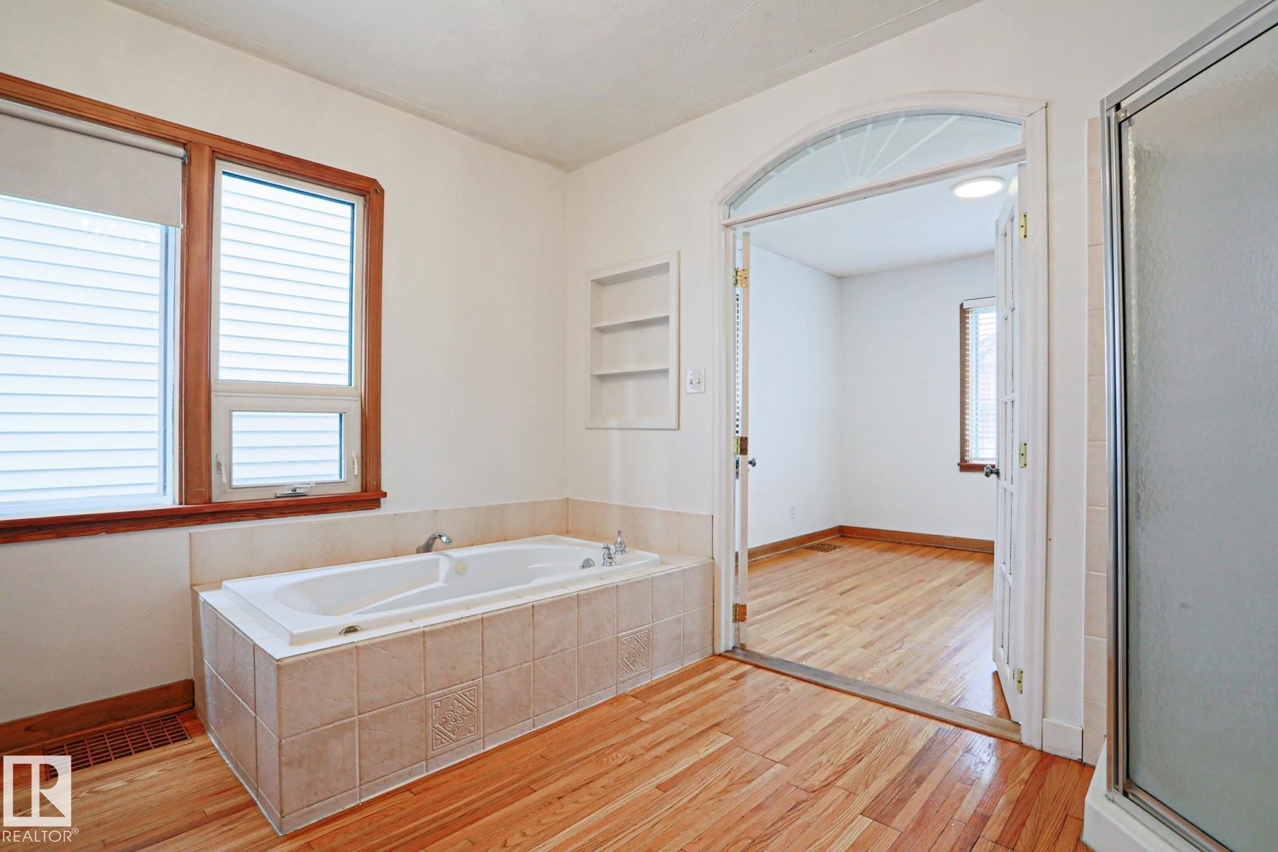 Jetted tub with tiled surround, wood-finish flooring, two windows with wood trim, built-in shelving, and an arched doorway with French doors - 9611 81 Avenue, Edmonton, AB - Indoor Photo Showing Bathroom