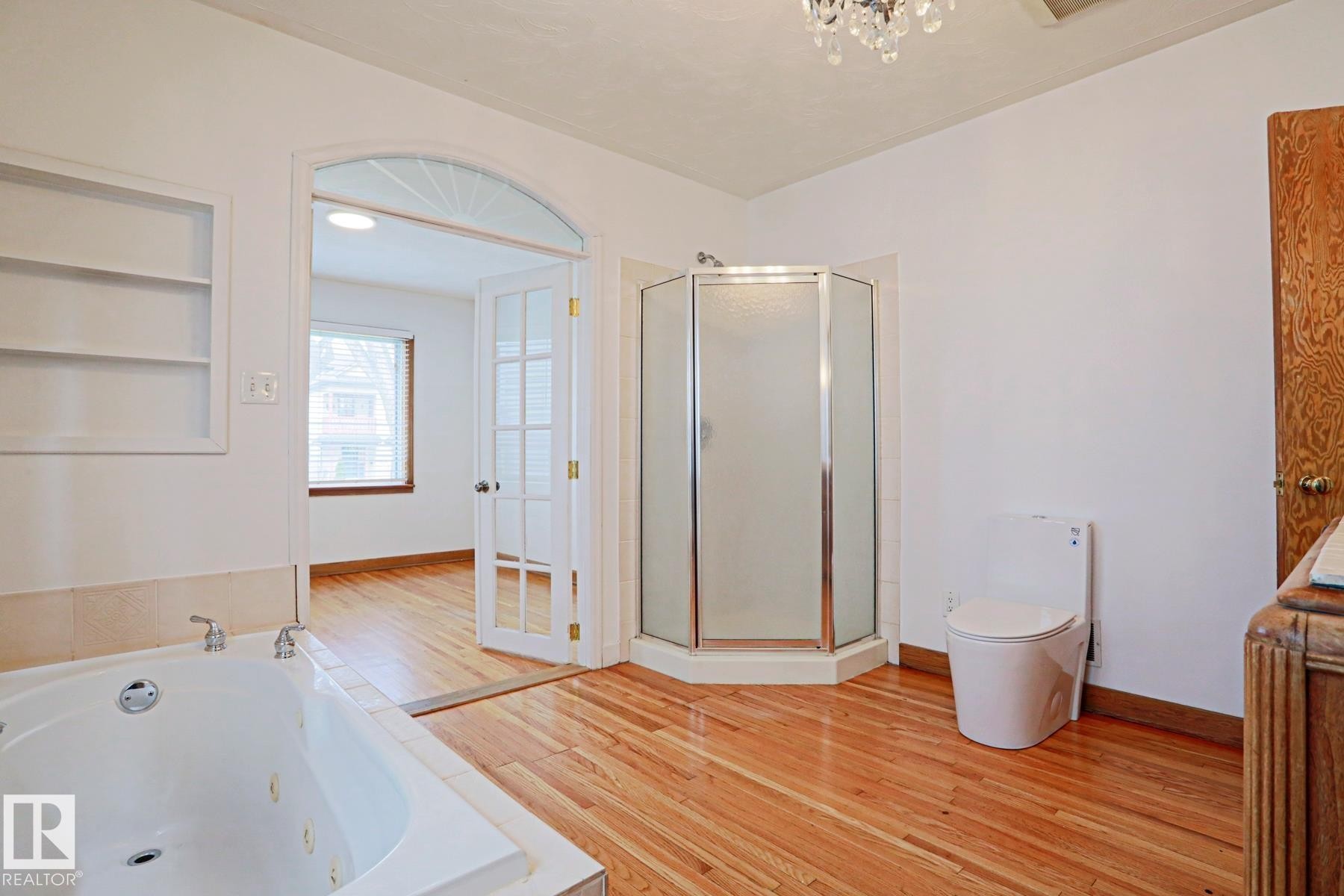 Bathroom featuring wood-finish flooring, a jetted soaking tub, a stand-up shower with frosted glass, and an arched entryway with French doors - 9611 81 Avenue, Edmonton, AB - Indoor