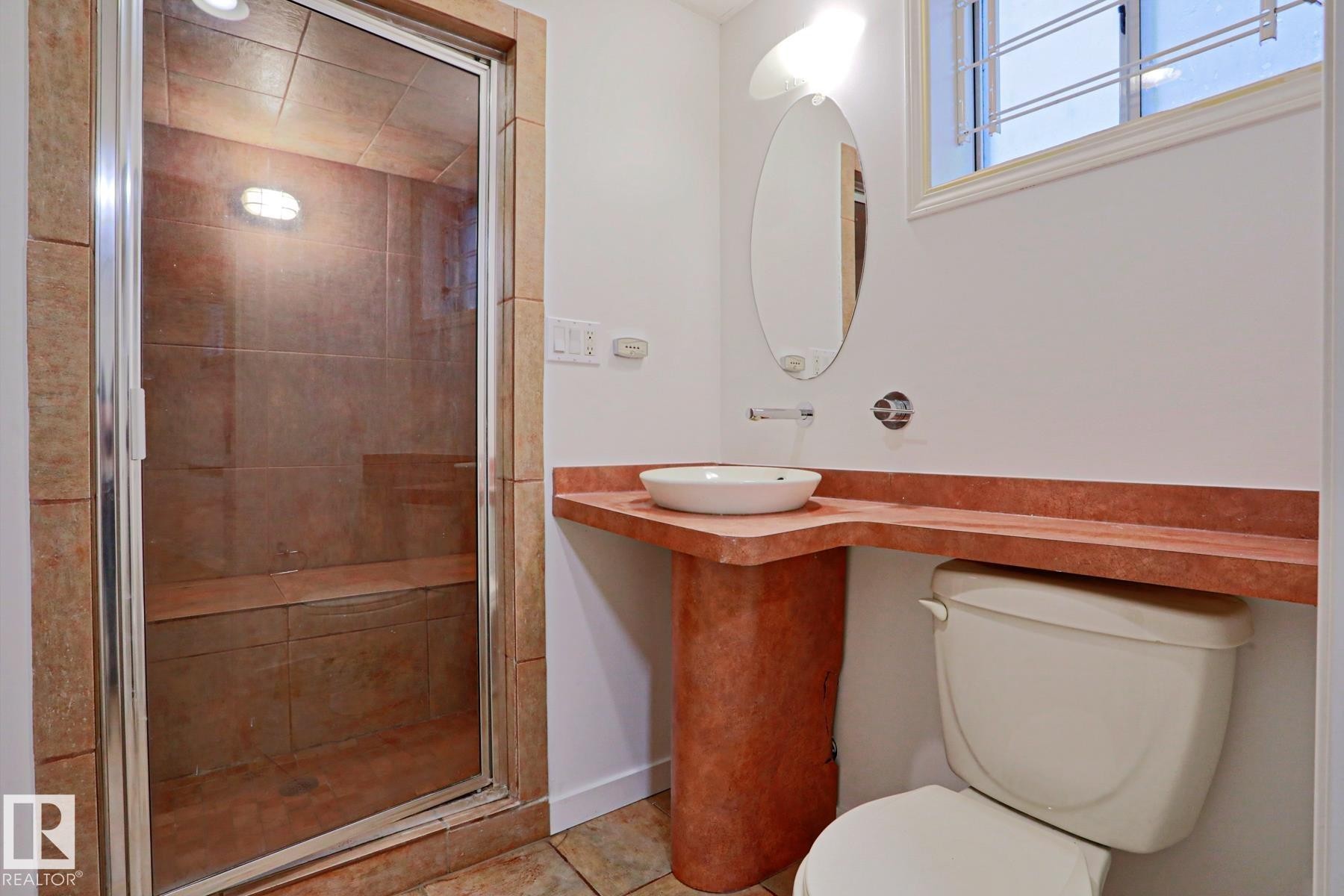 Bathroom featuring a tiled shower enclosure with a built-in bench, a vessel sink on a red-toned countertop, a wall-mounted mirror, and a window with security bars - 9611 81 Avenue, Edmonton, AB - Indoor Photo Showing Bathroom
