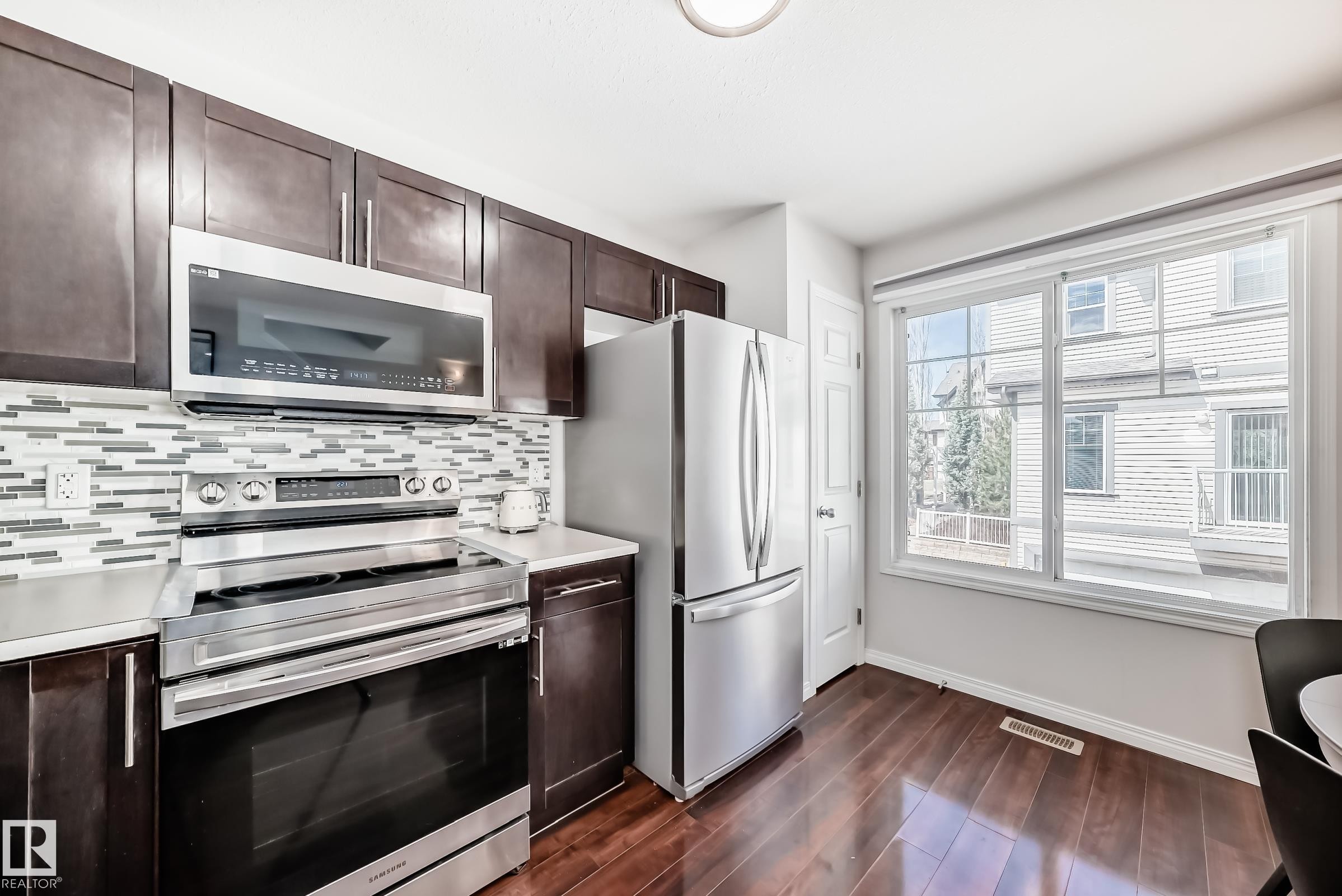 Kitchen featuring dark wood-finish cabinetry, stainless steel appliances, a mosaic tile backsplash, white countertops, and wood-finish flooring - 45 4850 Terwillegar Common, Edmonton, AB - Indoor Photo Showing Kitchen