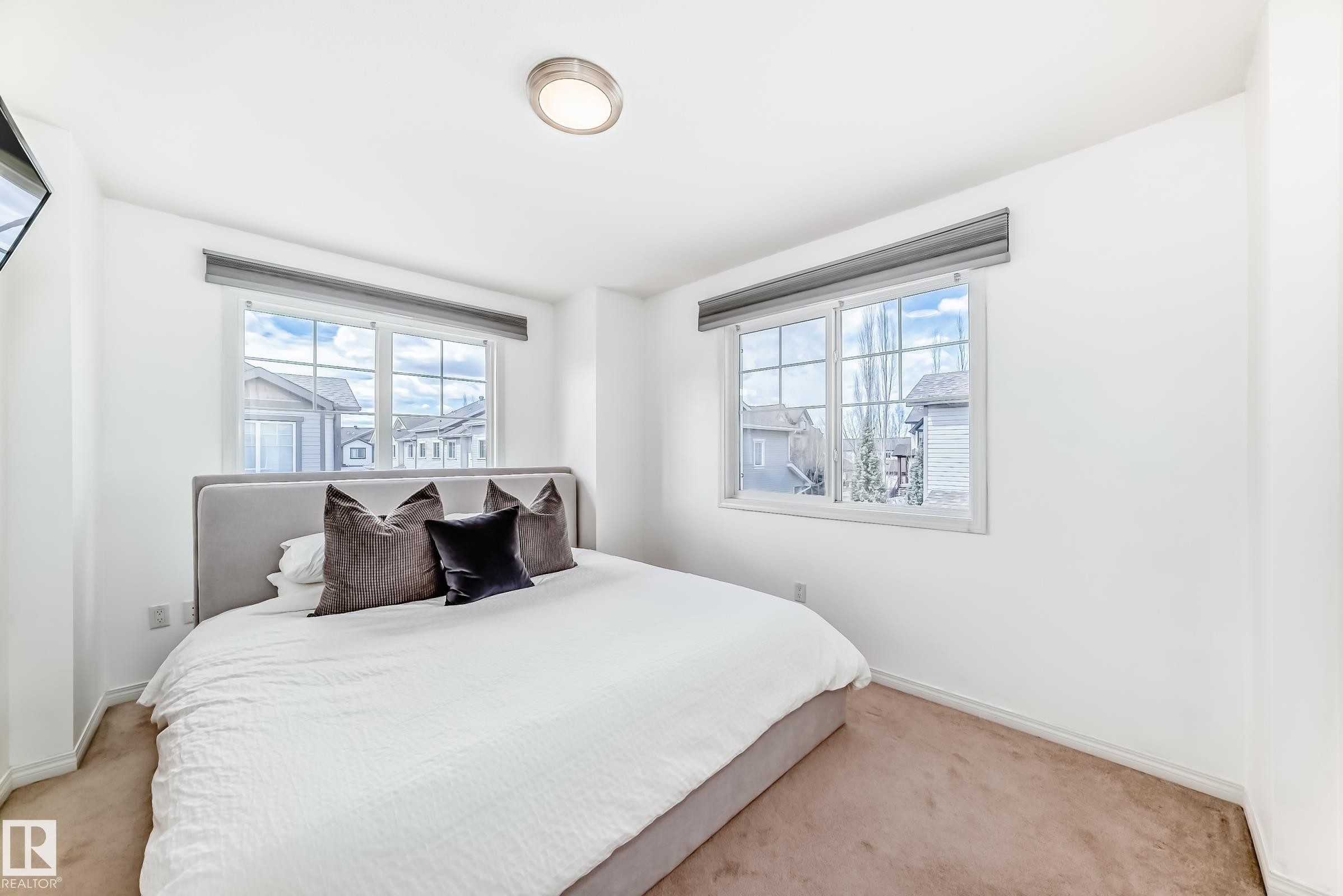 Bright bedroom featuring two large grid-pattern windows with roller shades, neutral carpeting, white walls, and a flush-mount ceiling light fixture - 45 4850 Terwillegar Common, Edmonton, AB - Indoor Photo Showing Bedroom