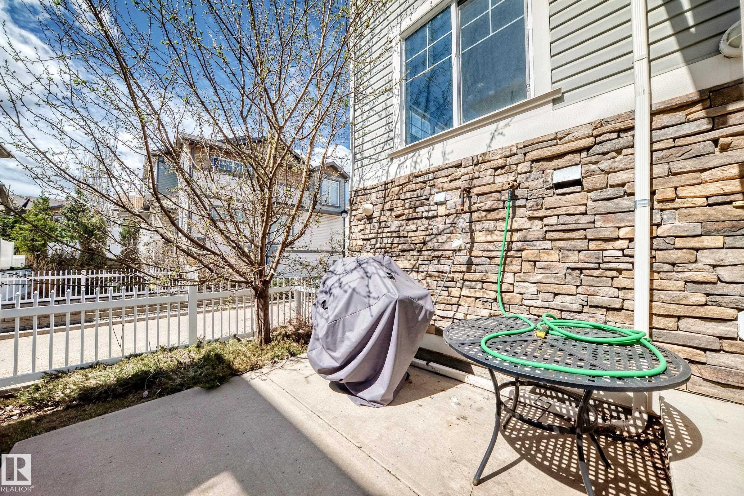 Concrete patio featuring a stone veneer exterior, a white picket fence, and a young deciduous tree - 45 4850 Terwillegar Common, Edmonton, AB - Outdoor
