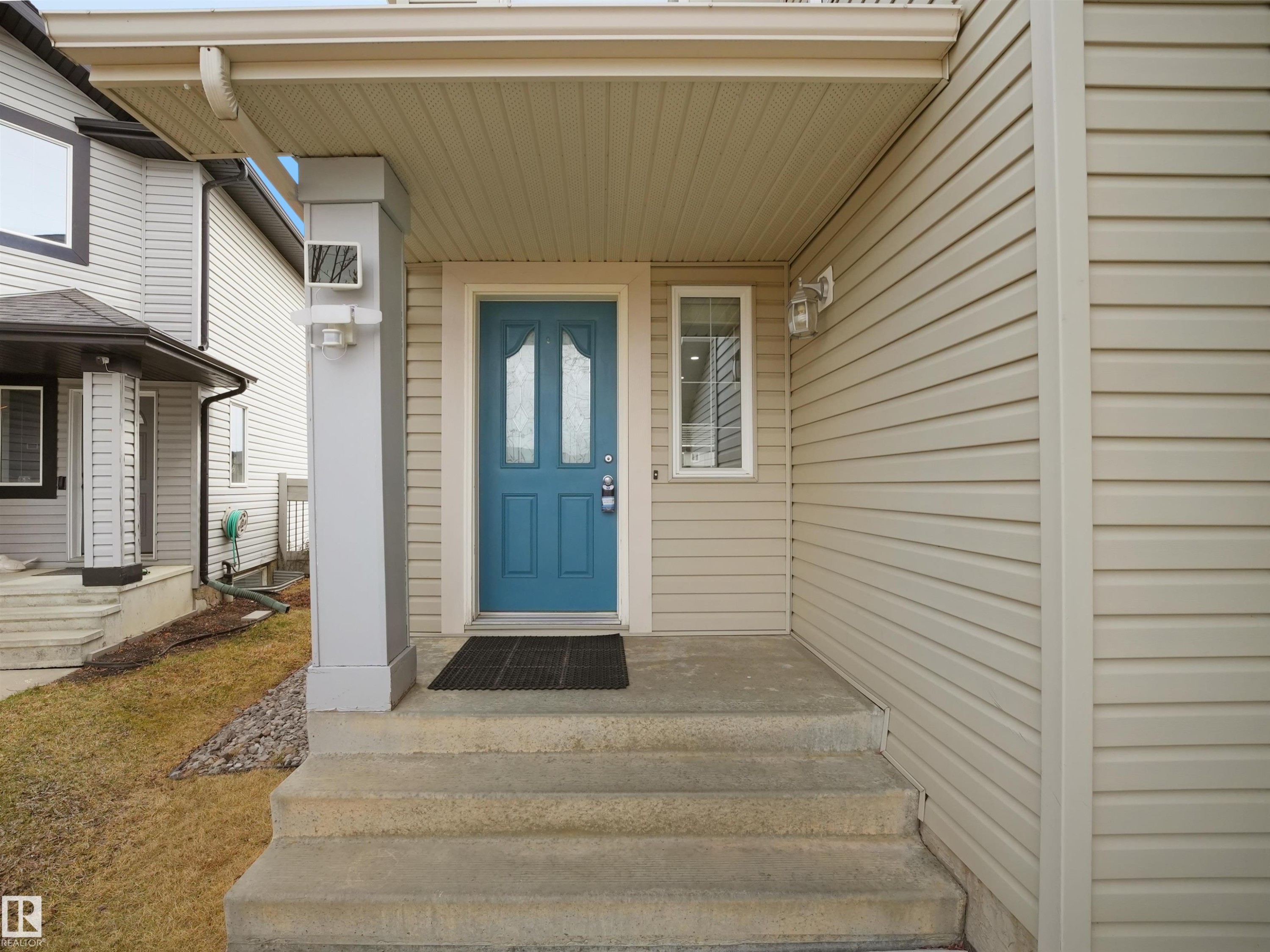 Inviting entryway featuring a teal panel door with decorative glass inserts, concrete steps, a covered porch, beige siding, and exterior lighting fixtures - 7407 Singer Landing, Edmonton, AB - Outdoor With Exterior