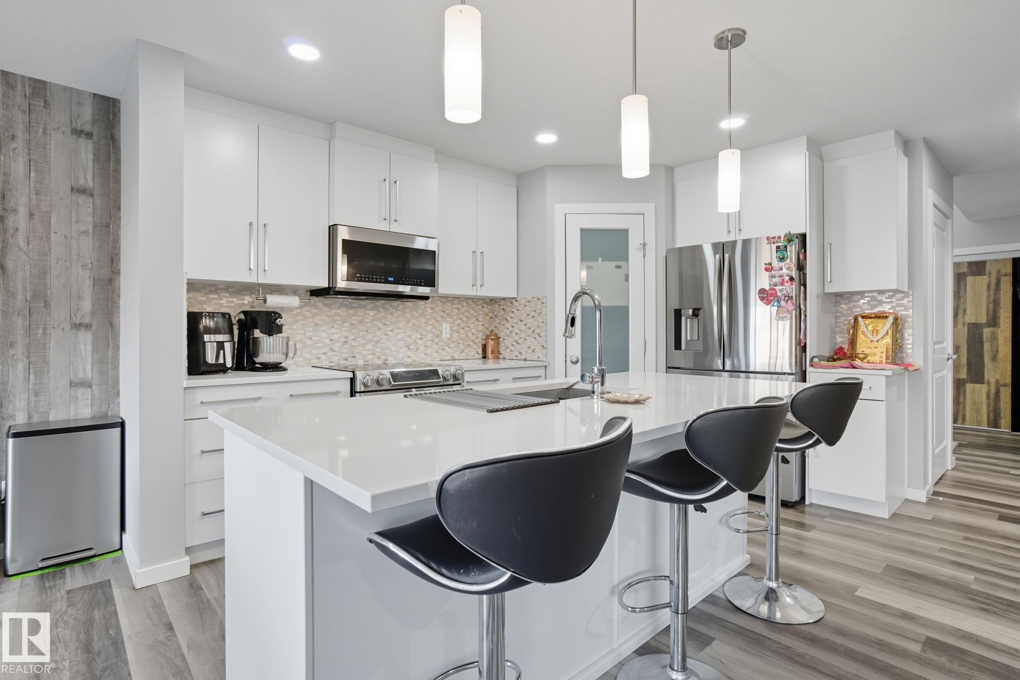 Modern kitchen featuring a central island with a solid surface countertop, white cabinetry, integrated stainless steel appliances, a mosaic tile backsplash, and wood-finish flooring - 2039 Wonnacott Way, Edmonton, AB
