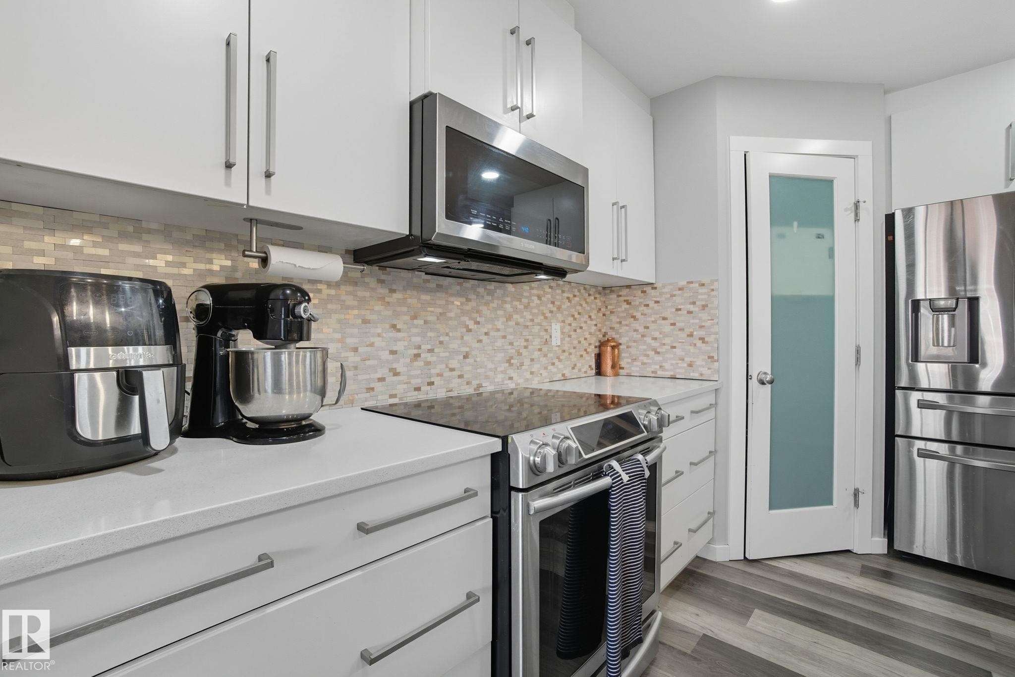 Modern kitchen featuring stainless steel appliances, white cabinetry, a mosaic tile backsplash, light-toned countertops, and wood-finish flooring - 2039 Wonnacott Way, Edmonton, AB