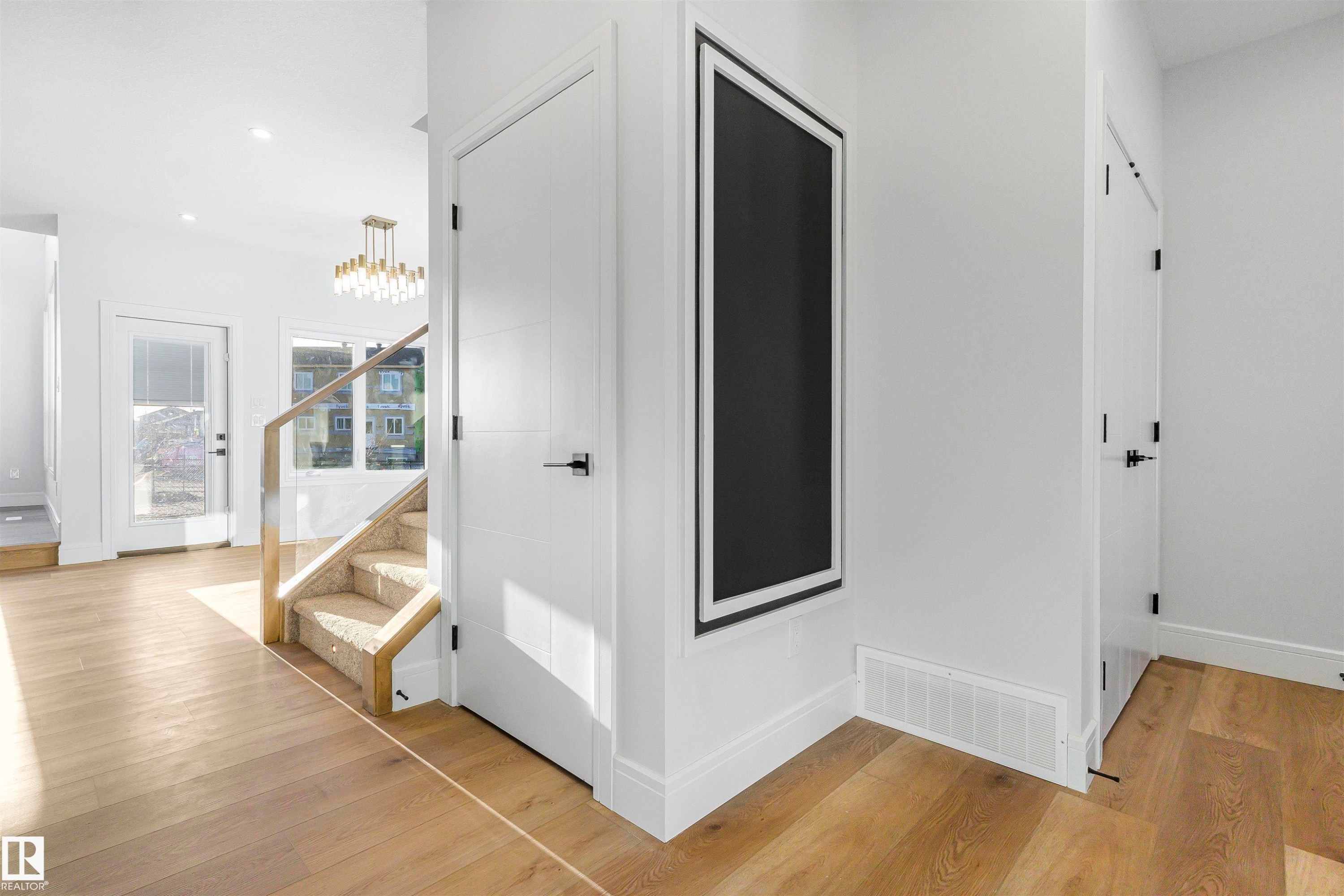 Hardwood-finish flooring throughout, featuring a staircase with glass panels and a carpeted tread, a modern chandelier, and white paneled doors with matte black hardware - 372 Creekside Green, Leduc, AB - Indoor Photo Showing Other Room