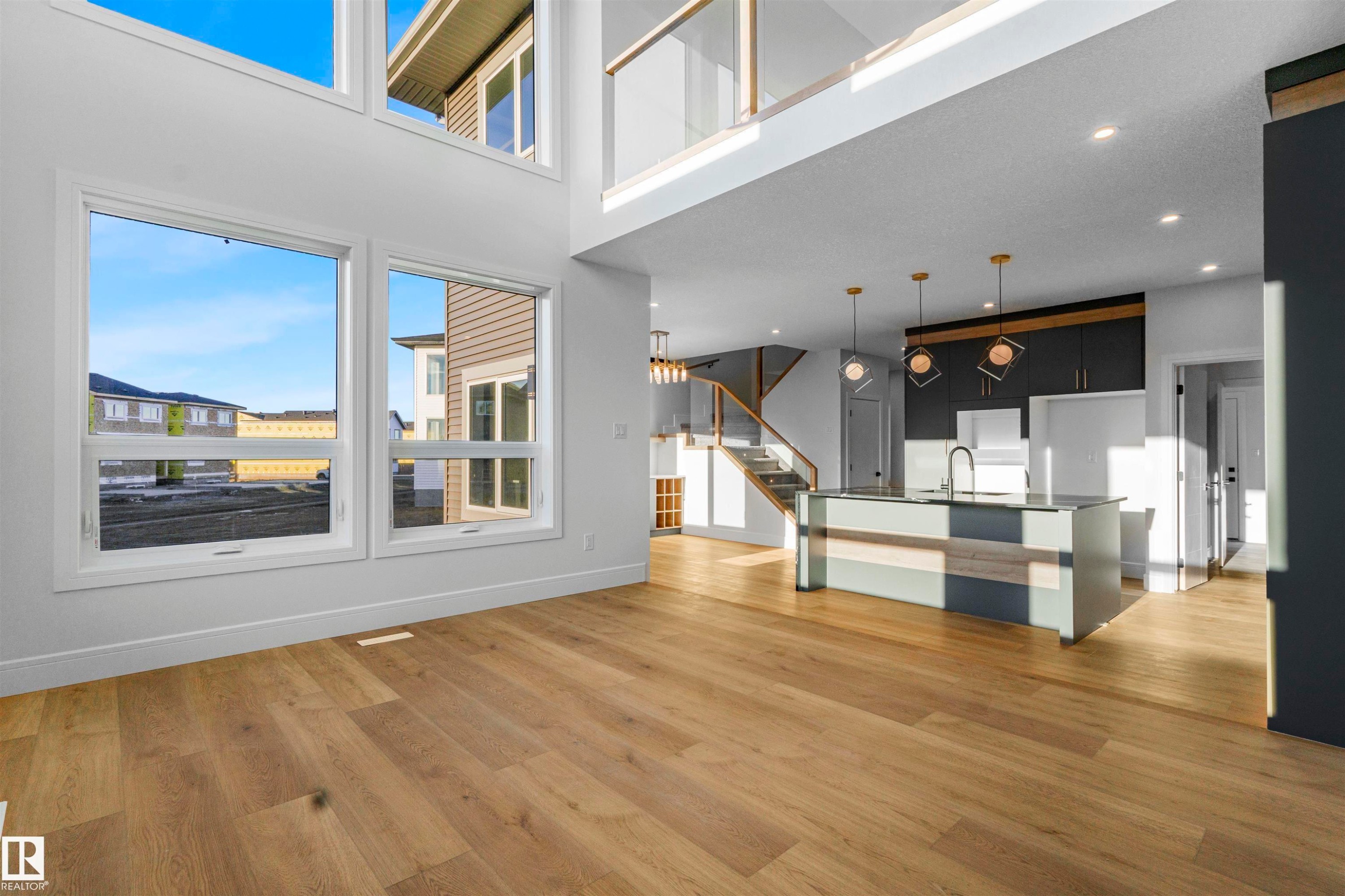 Open-concept living space featuring high ceilings, wood-finish flooring, a contemporary kitchen with an island, and a prominent staircase with glass railing - 372 Creekside Green, Leduc, AB - Indoor