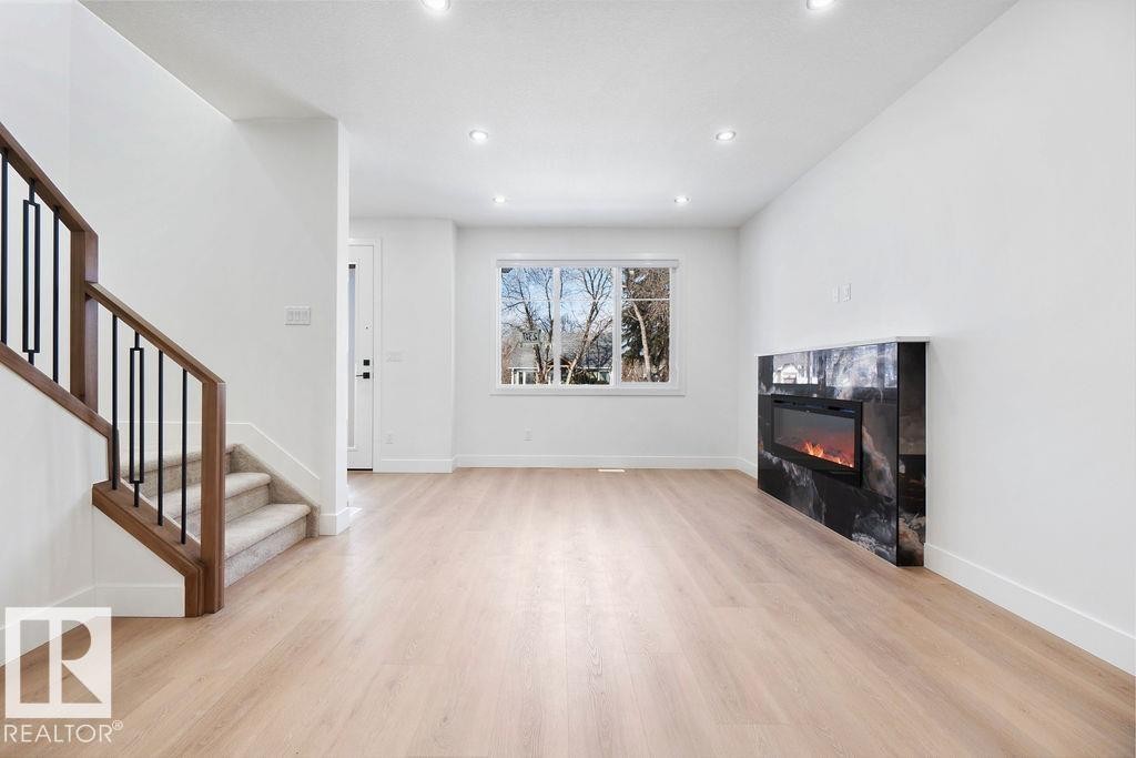 The interior features light-colored flooring, a staircase with a wooden handrail and dark balusters, and a contemporary fireplace with a dark, veined surround - 12307 91 St Nw, Edmonton, AB - Indoor Photo Showing Other Room With Fireplace