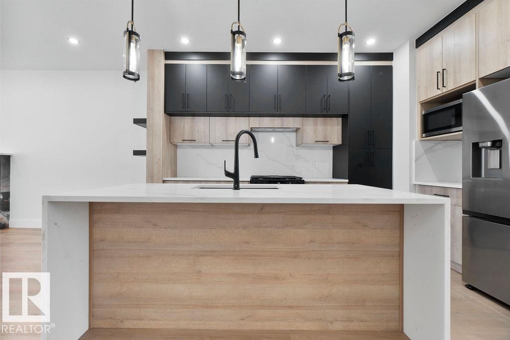 Modern kitchen featuring a large island with a light wood panel accent and a white countertop, dark and light wood cabinetry, and a stainless steel refrigerator - 12307 91 St Nw, Edmonton, AB - Indoor Photo Showing Kitchen With Upgraded Kitchen