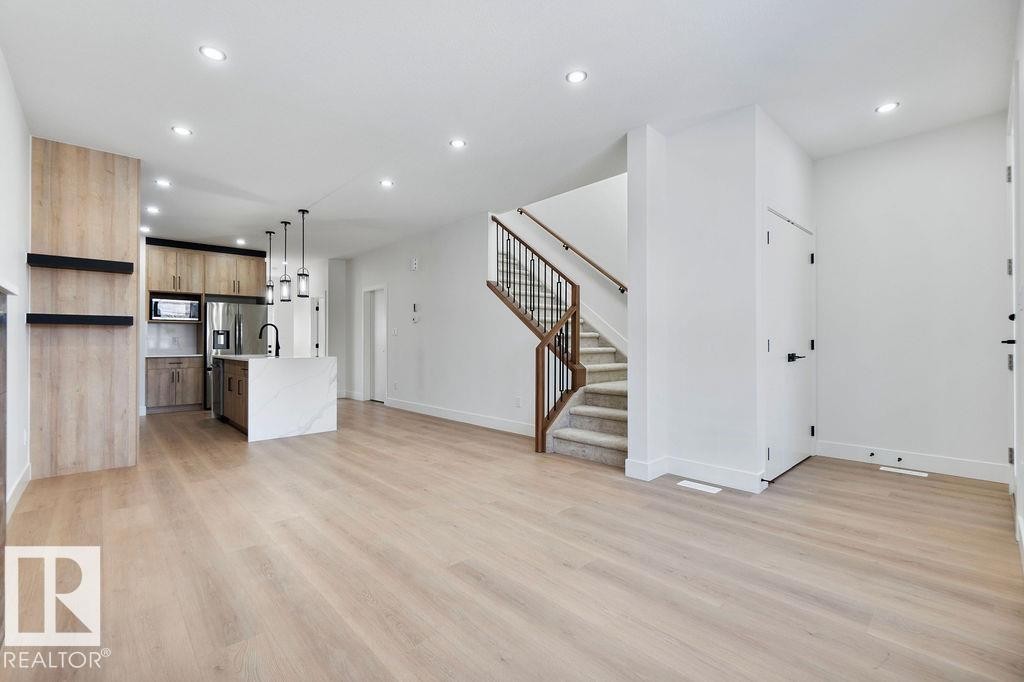 Open concept living space featuring light-colored flooring, a modern kitchen with a white island, and a staircase with a dark metal railing - 12307 91 St Nw, Edmonton, AB - Indoor Photo Showing Other Room