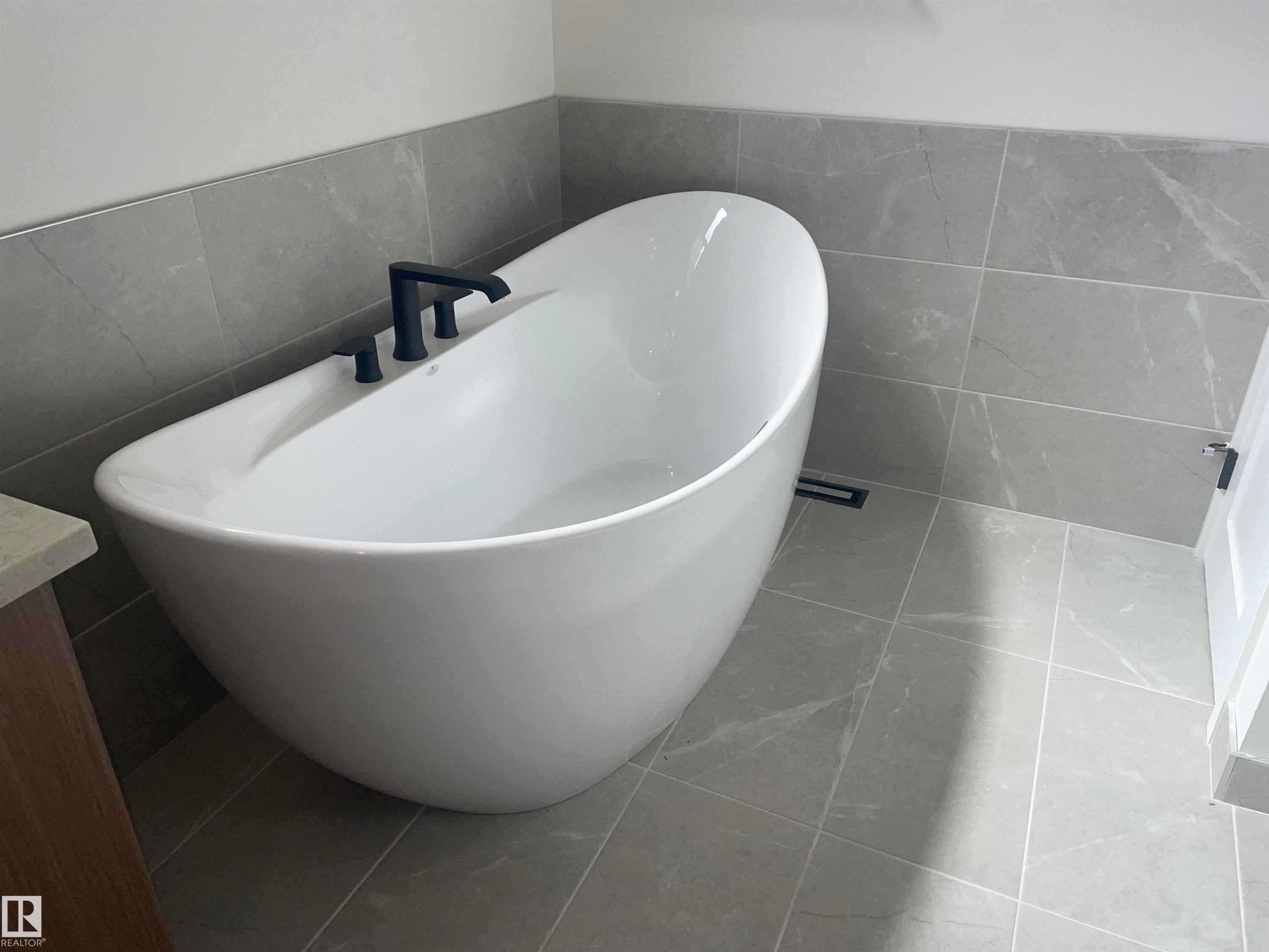 Freestanding soaking tub with matte black fixtures, set against light gray tile flooring and wainscoting - 14344 92A Avenue, Edmonton, AB - Indoor Photo Showing Bathroom