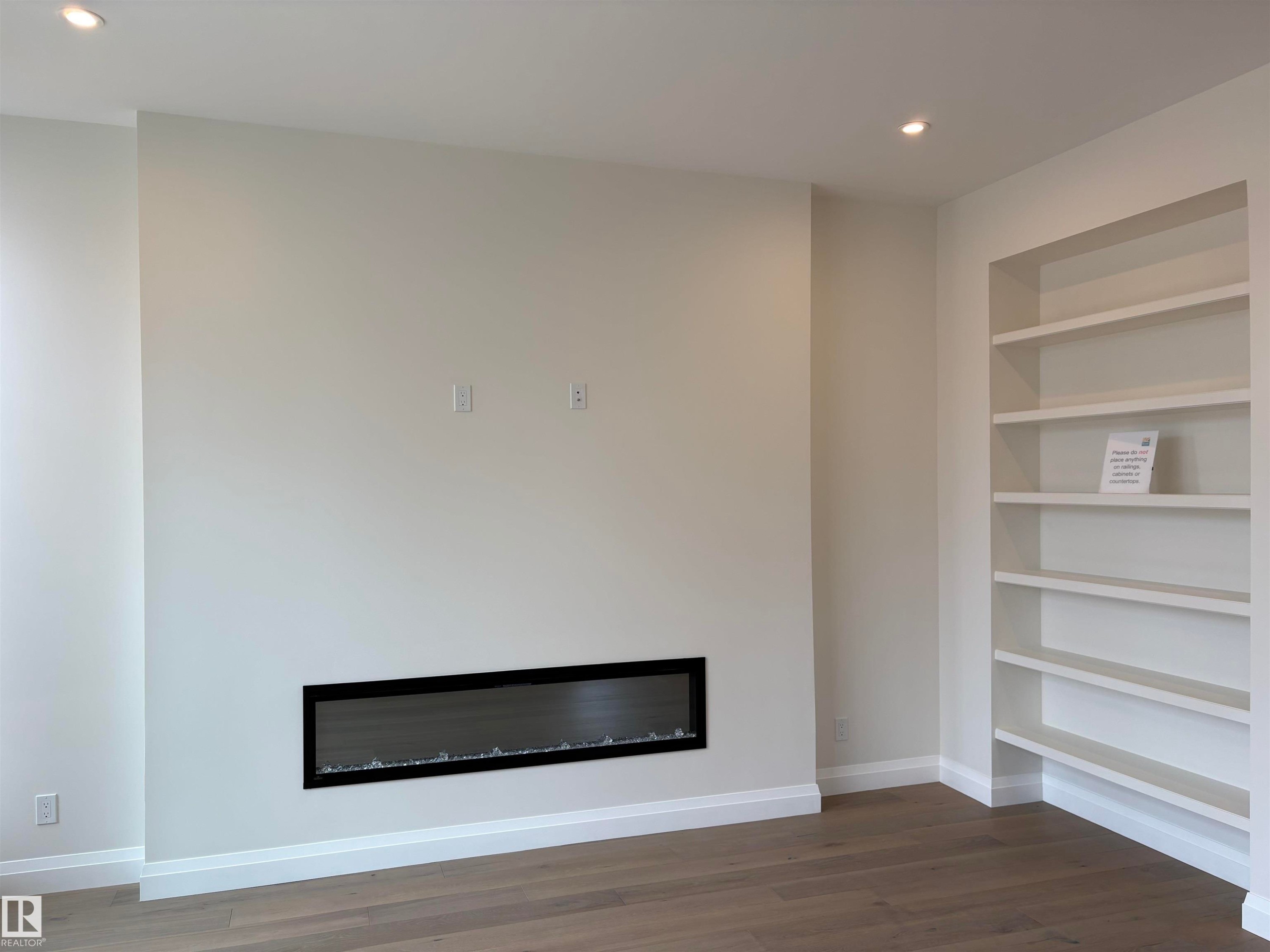 Linear electric fireplace with a black frame, recessed built-in shelving, wood-finish flooring, white baseboards, and neutral wall paint - 14344 92A Avenue, Edmonton, AB - Indoor With Fireplace