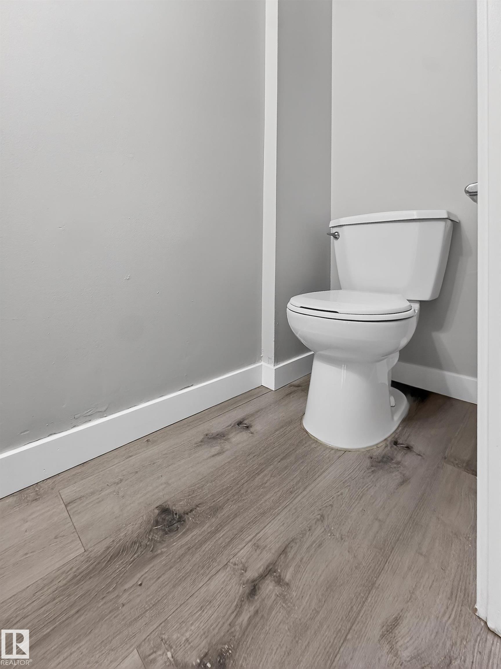 White toilet with a chrome flush handle, set against gray walls with white baseboards and wood-finish flooring - 18 Lakewood Village, Edmonton, AB - Indoor Photo Showing Bathroom