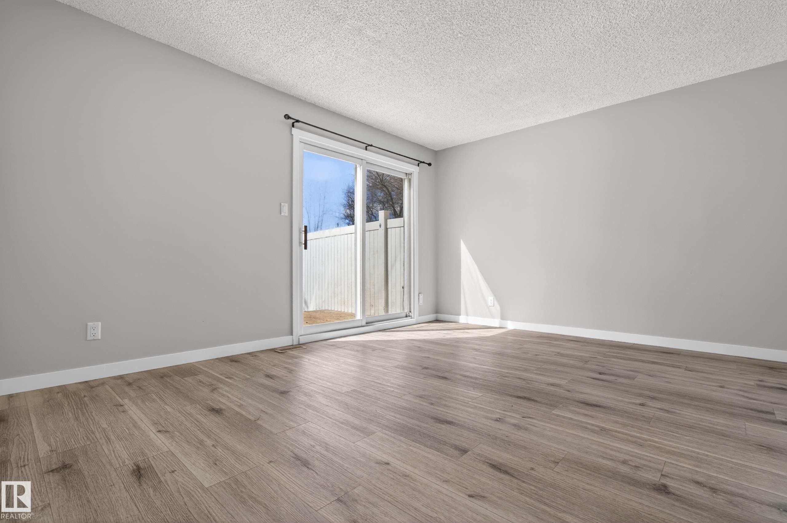 Spacious room featuring wood-finish flooring, light grey painted walls, white baseboards, and a sliding glass door providing access to a private outdoor area with a white fence - 18 Lakewood Village, Edmonton, AB - Indoor Photo Showing Other Room