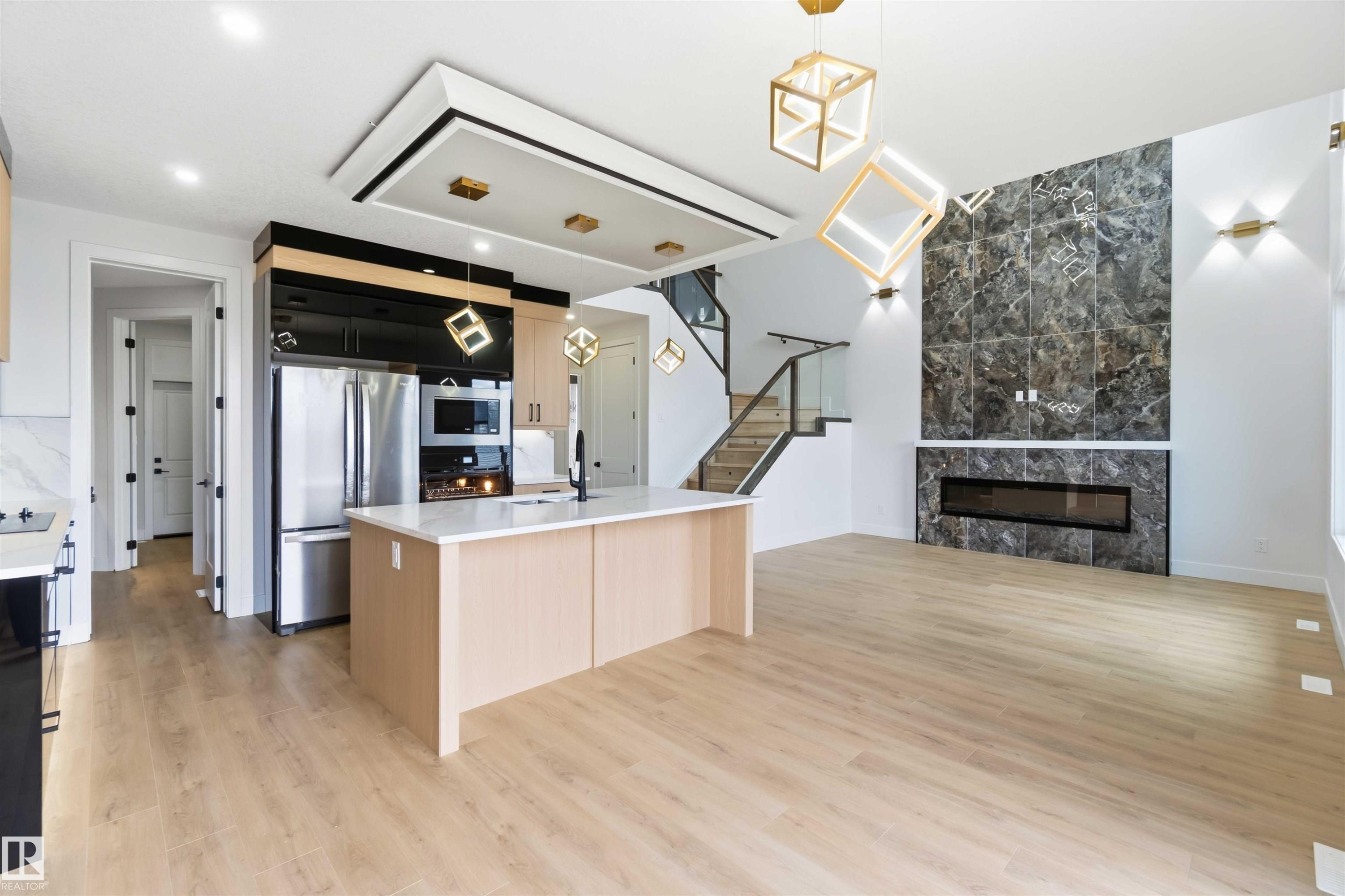 Open-concept living space featuring light wood-finish flooring, a contemporary kitchen island with a white countertop, and a striking fireplace wall with large-format dark stone-look tiles - 417 Pine Point(E), Leduc, AB - Indoor Photo Showing Kitchen With Fireplace