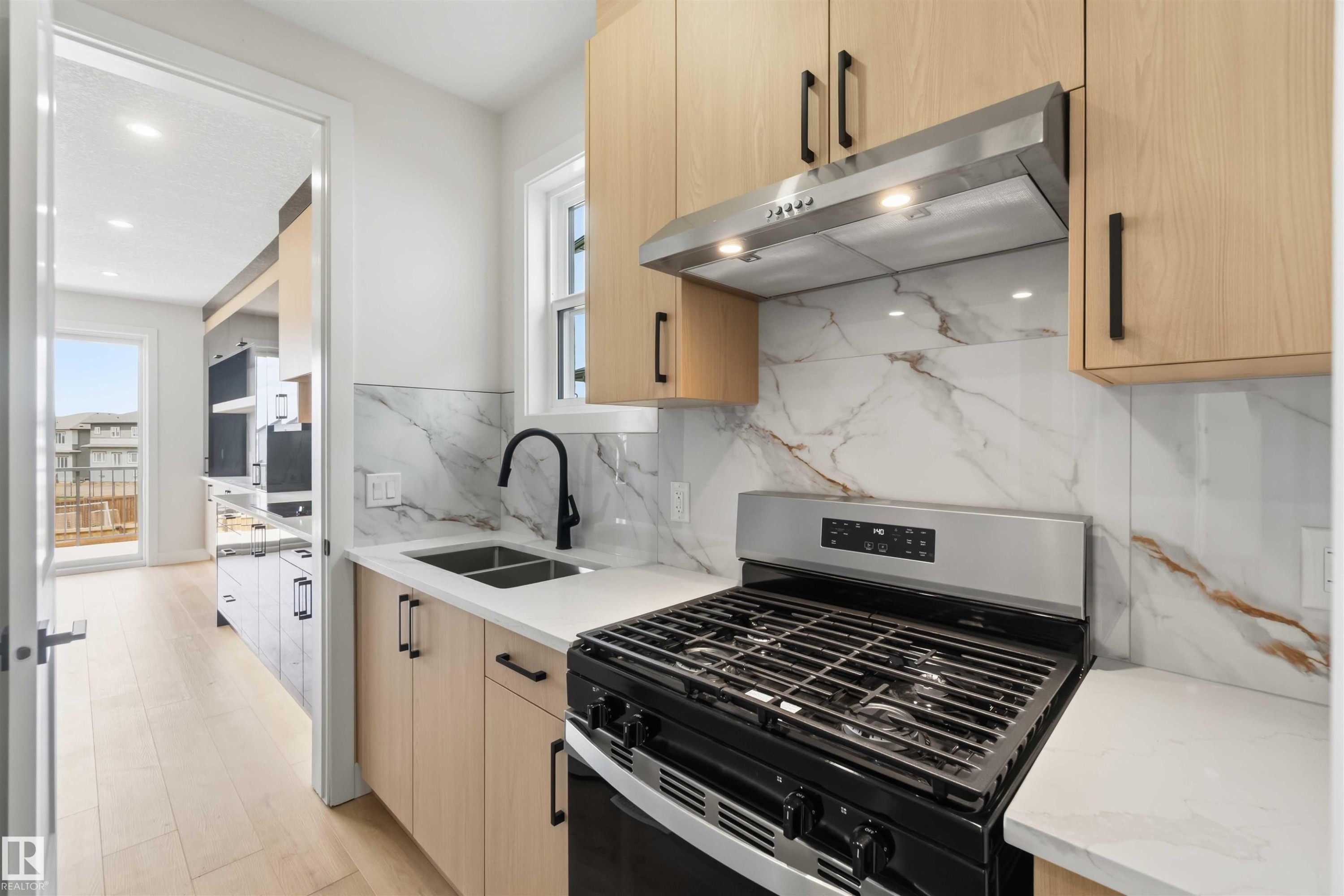 Modern kitchen featuring light wood-finish cabinetry, a stainless steel range with overhead ventilation, a dual basin sink with a matte black faucet, and a patterned backsplash with white countertops - 417 Pine Point(E), Leduc, AB - Indoor Photo Showing Kitchen With Double Sink With Upgraded Kitchen