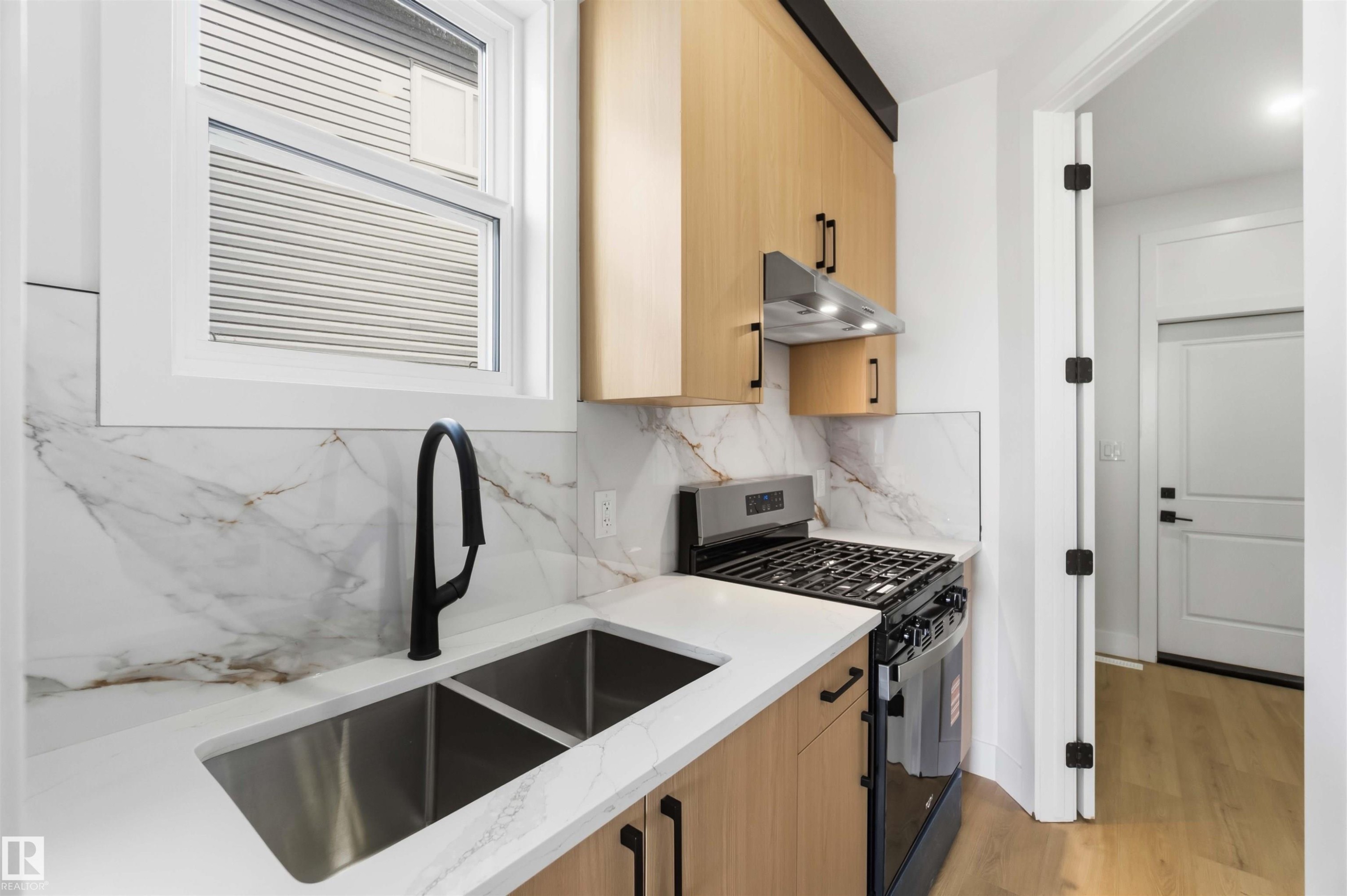 Modern kitchen featuring a stainless steel double sink with a matte black faucet, light wood-finish cabinetry, white countertops, and a full tile backsplash - 417 Pine Point(E), Leduc, AB - Indoor Photo Showing Kitchen With Double Sink