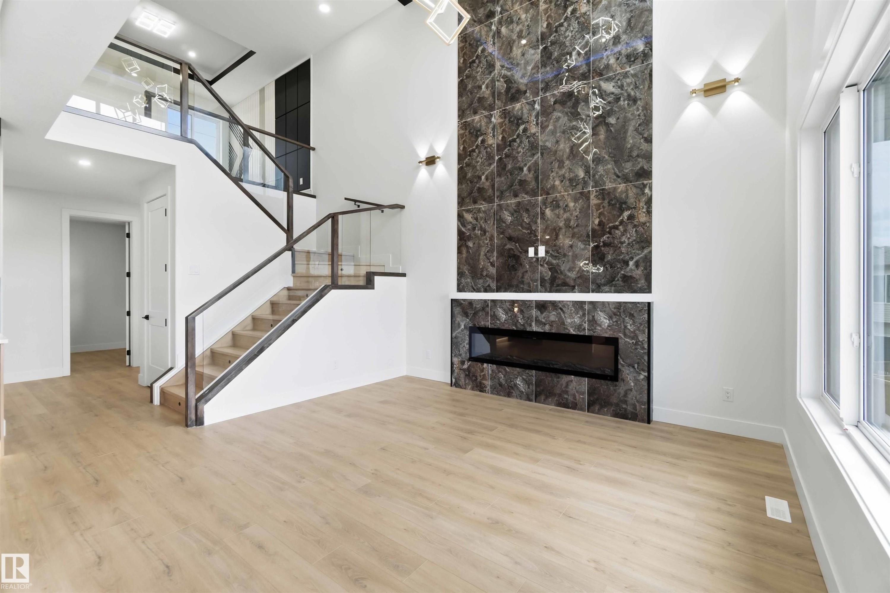 Two-story living area featuring wood-finish flooring, a contemporary linear fireplace with a dark marble-patterned surround, and a modern staircase with glass panel railings - 417 Pine Point(E), Leduc, AB - Indoor With Fireplace
