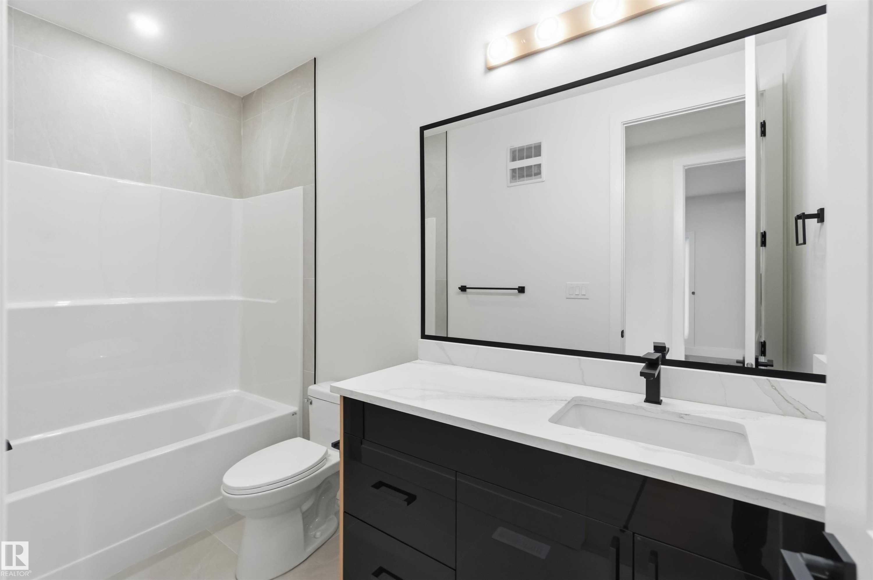 Bathroom featuring a white tub/shower combination with light-colored tile surround, a toilet, a dark vanity with a white countertop, and a large mirror with a matte black frame - 417 Pine Point(E), Leduc, AB - Indoor Photo Showing Bathroom