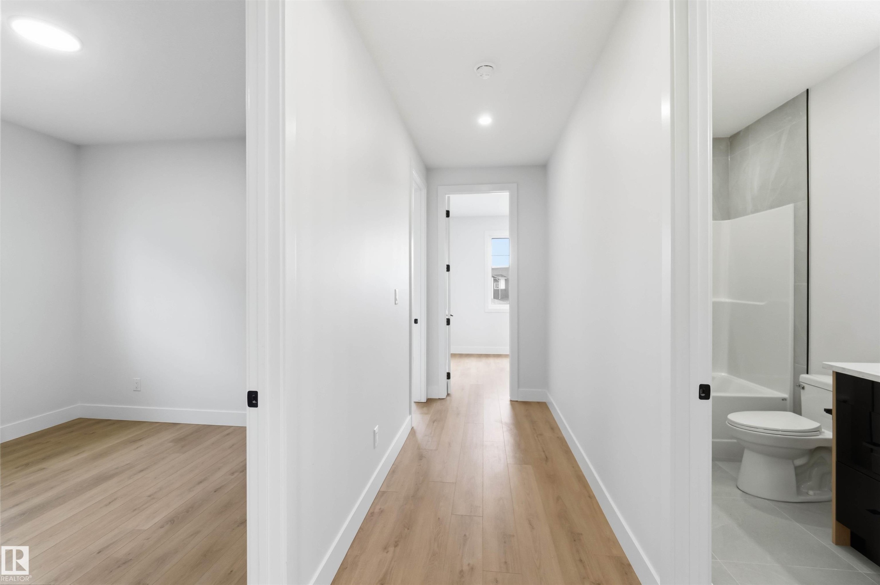 Clean white walls and wood-finish flooring characterize this hallway, offering access to multiple rooms including a bathroom with a white tub and toilet, and an additional room with a recessed ceiling light - 417 Pine Point(E), Leduc, AB - Indoor Photo Showing Other Room